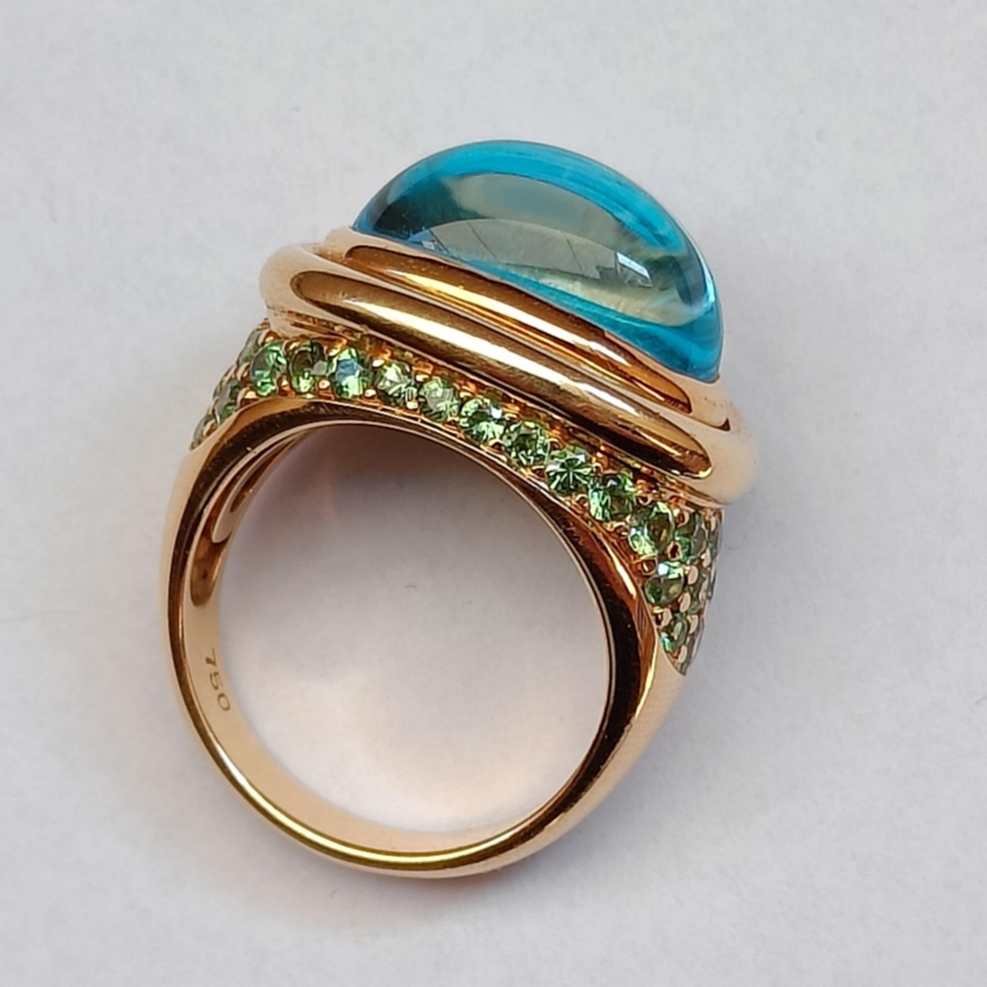 A large 18k yellow gold ring adorned with a blue topaz oval cabochon surrounded by a pavé of tsavorites (green garnets)

Stamped 750. There is another stamp on the external shank of the ring, but it's very small and I couldn't identify it.

Size: FR