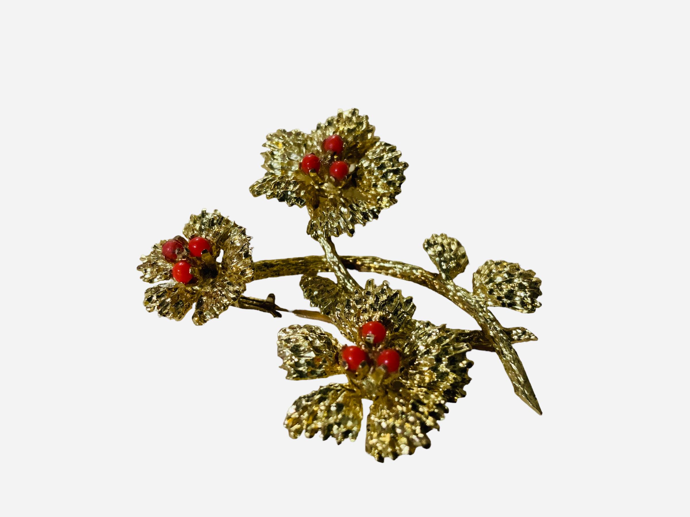This is an 18K Gold and Coral Brooch. It depicts a yellow gold two branches with leaves and three flowers brooch. Each one of the flowers has five petals and are adorned with three coral beads. The brooch has a trombone clasp. It is hallmarked 18k.