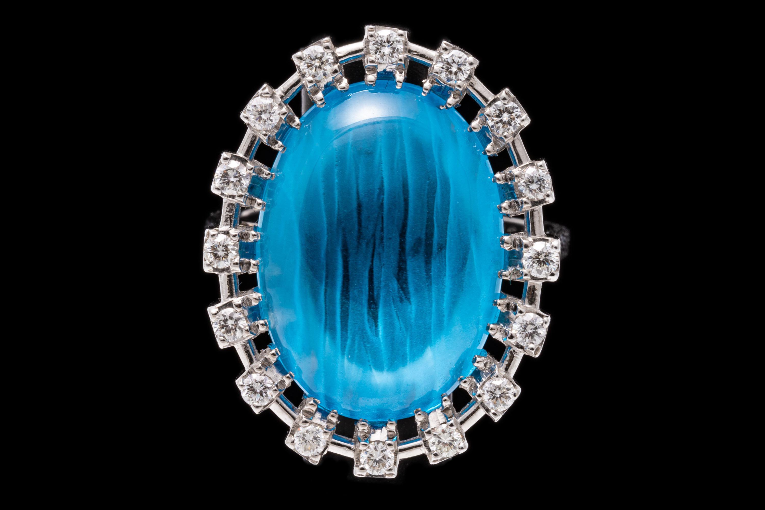 Bague en or blanc 18k. Cette magnifique bague de cocktail est sertie au centre d'une topaze bleue de couleur bleu moyen, de taille ovale fantaisie, entourée d'un halo contemporain de diamants ronds à facettes, d'une valeur approximative de 0,16 TCW,