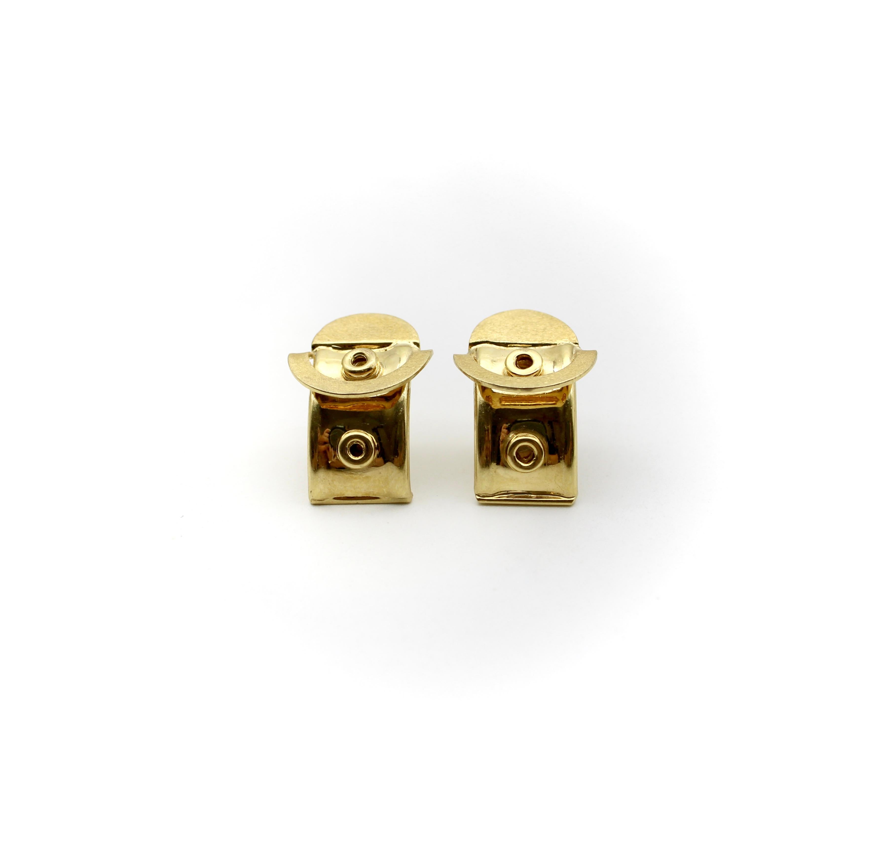 These 18k gold earrings appear to be “buckled” into your earlobe. A circle at the top of the earring alludes to a buckle that curves and flows under the ears in huggie fashion. The gold has both shiny and matte surfaces, with raised circular