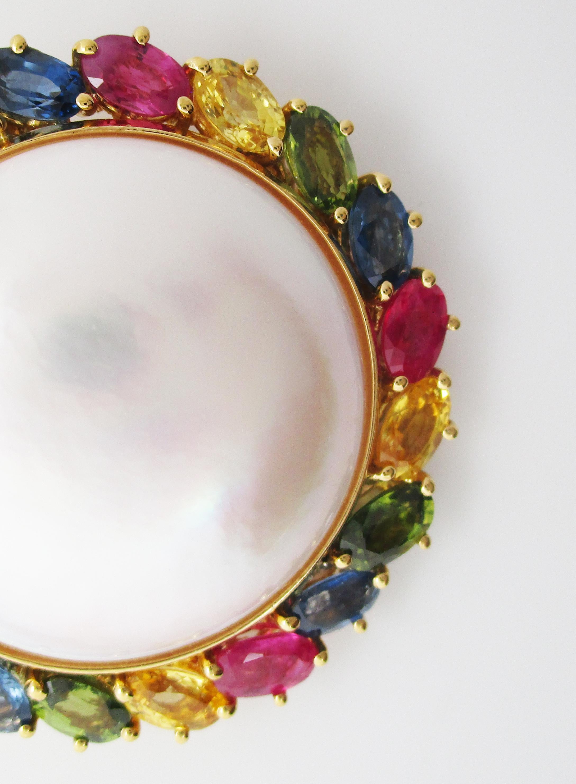 This stunning brooch is in 18k yellow gold and features an impressively huge mabe pearl center surrounded by a frame of multi-colored oval cut sapphires ranging in color from red to blue, green, and yellow. The rainbow iridescence of the pearl