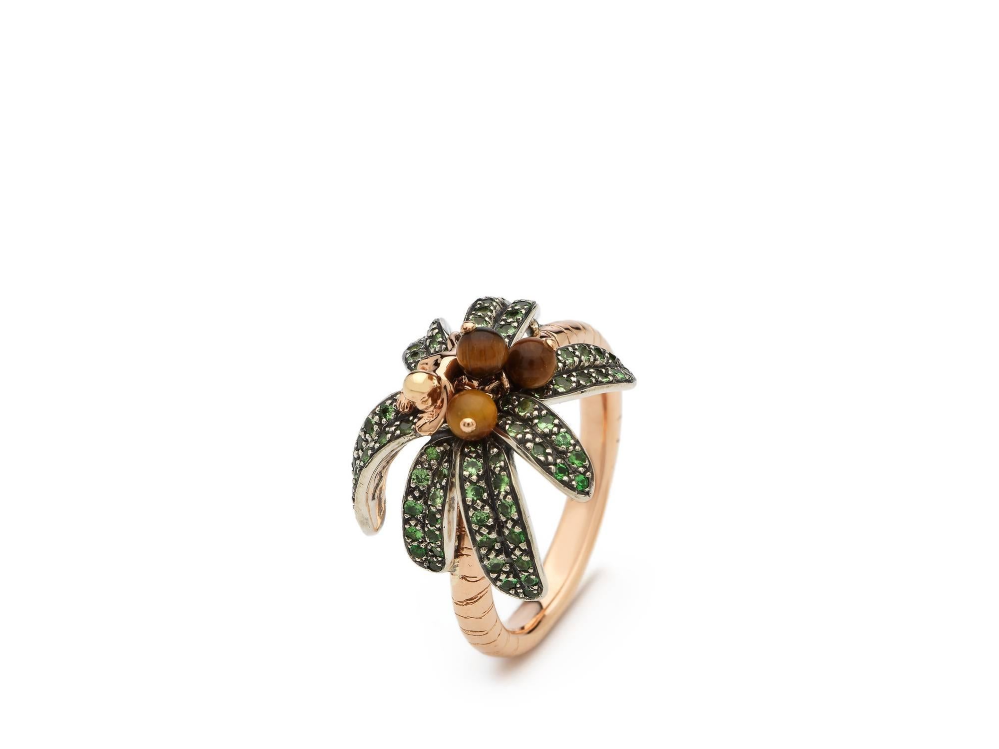 A baby monkey crafted from 18k rose gold clambers amid palm leaves embellished with green tsavorites on this 18k rose gold ring. With its band textured to evoke a palm tree’s trunk, tiger’s eye coconuts are also set on the ring, that gently move as