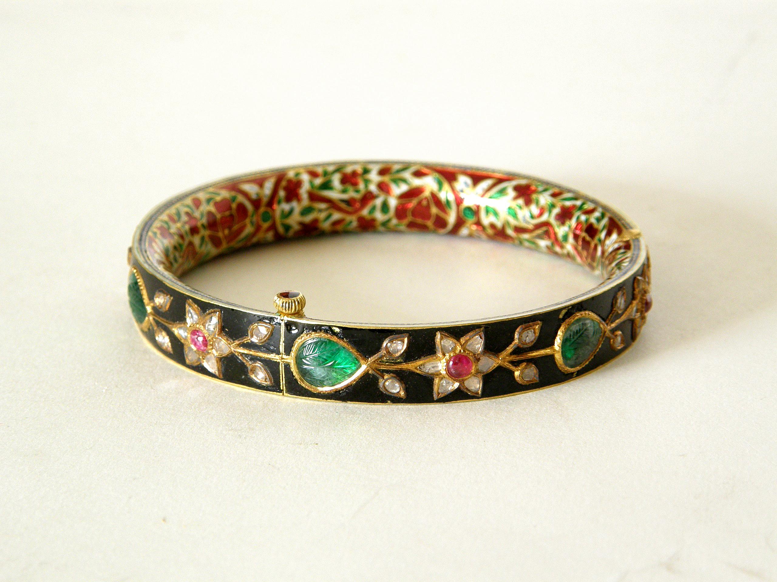 This Mughal style hinged bracelet is made of 18K gold. The outside of the bangle has a rich, black enamel that beautifully offsets the abstract floral design set with diamonds, rubies, and pear-shaped emeralds. The interior of the bangle has an
