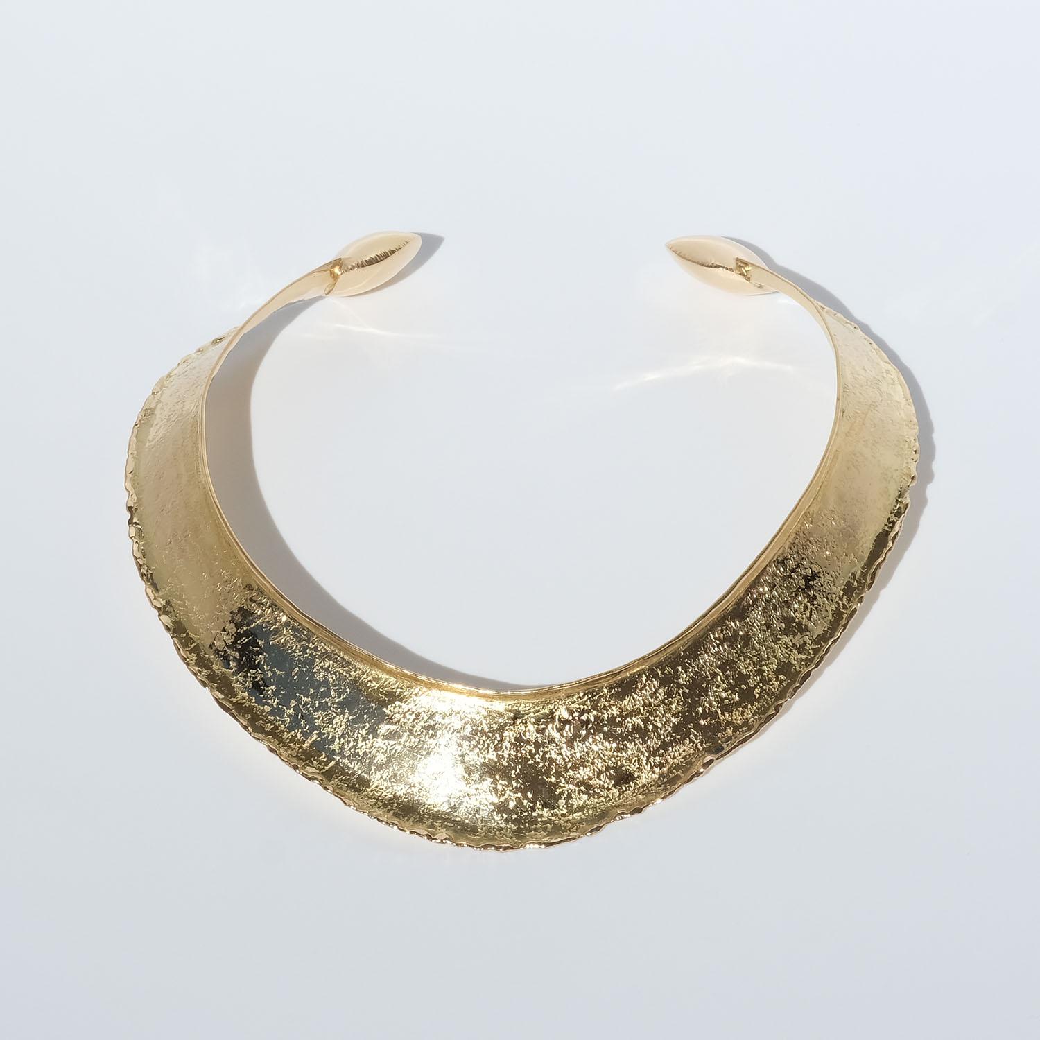 This 18 karat gold neck ring has a shiny, textured surface. The shape of the neck ring may be described as semi oval with an accentuated front. The neck ring's arms are equipped with square-shaped golden pillows on its ends.

The neck ring looks