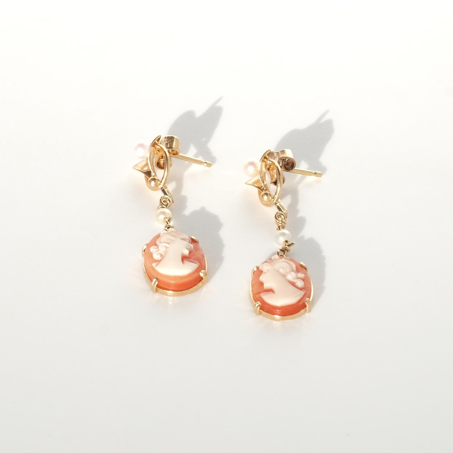 Questi orecchini vintage sono realizzati in oro 18 carati e progettati come delicati perni pendenti. Ogni orecchino presenta un cammeo di conchiglia intagliato che raffigura un profilo femminile classico, con la figura bianco crema che si staglia su