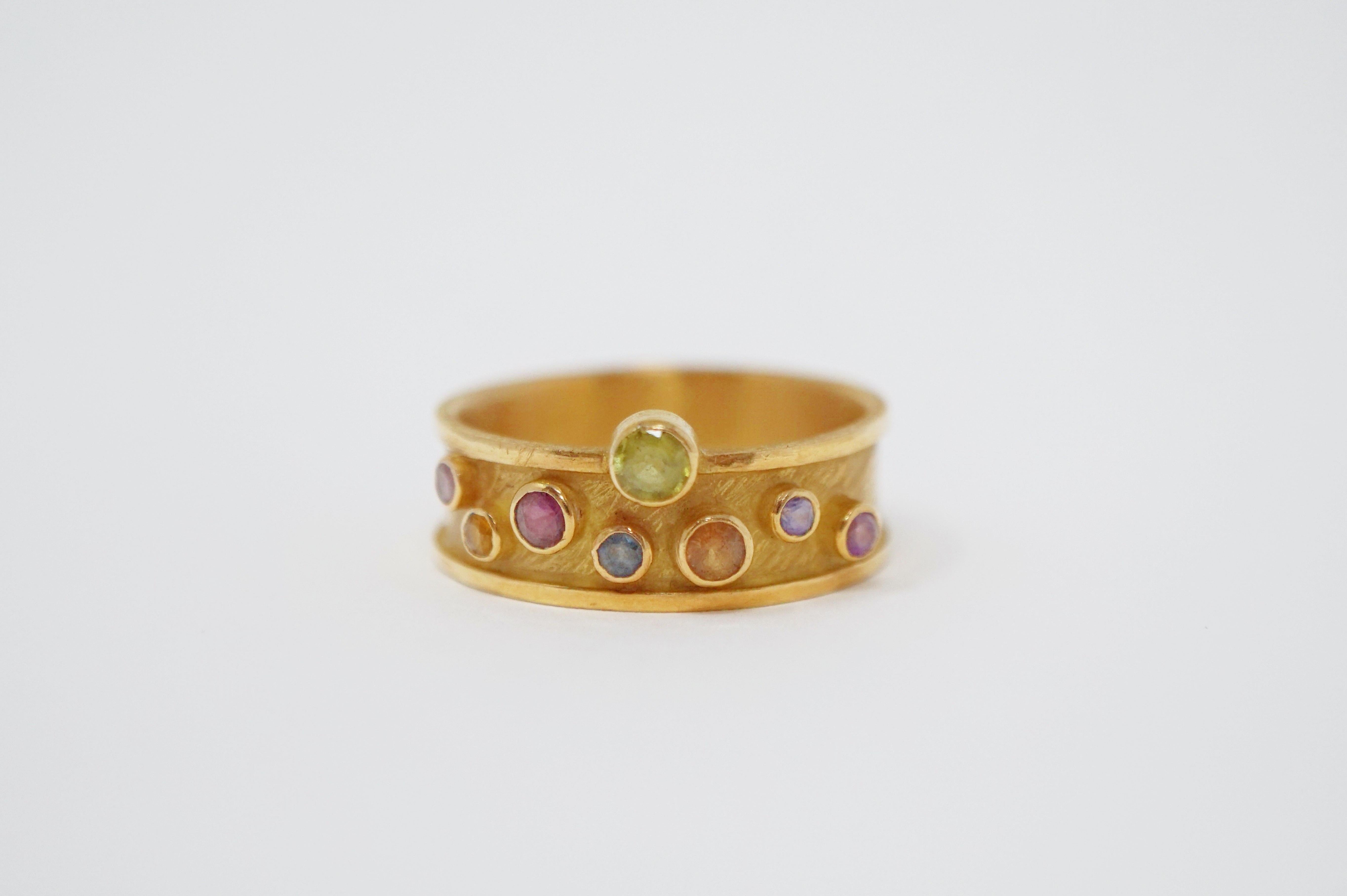 This gorgeous 18 karat yellow gold band ring by Barbara Heinrich is a highly wearable work of art, featuring eight round faceted bezel set sapphires in a rainbow of shades including ruby, amber, cornflower, light green and purple. The textured band