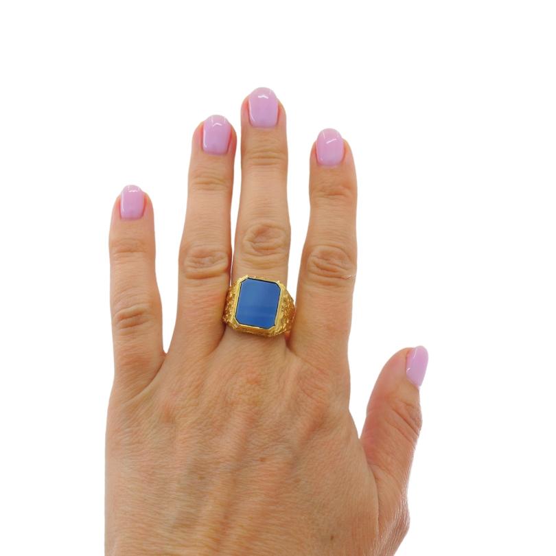 A chunky 18k gold signet ring, featuring blue agate.
The face of the ring is set with a rectangular stone. It’s cut with smooth edges and set flush in the gold frame. The gold has a textured finish with a rugged pattern along the shoulders. These