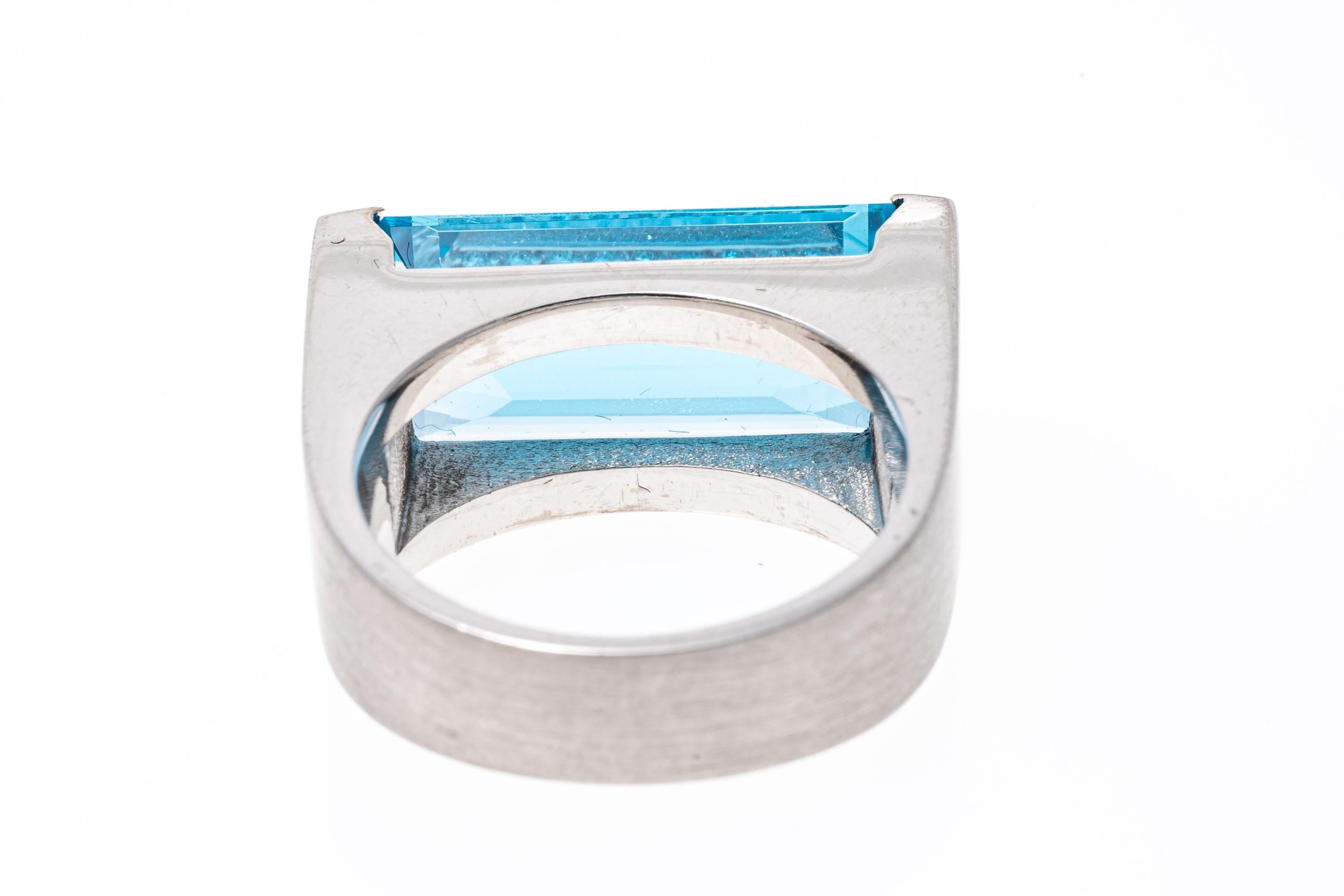 18k white gold ring. This stunning, white gold ultra modern ring has an elongated, narrow rectangular faceted, medium to light blue color blue topaz, approximately 9.74 CTS, half bezel set by wide, columnar sides, and decorated with round faceted