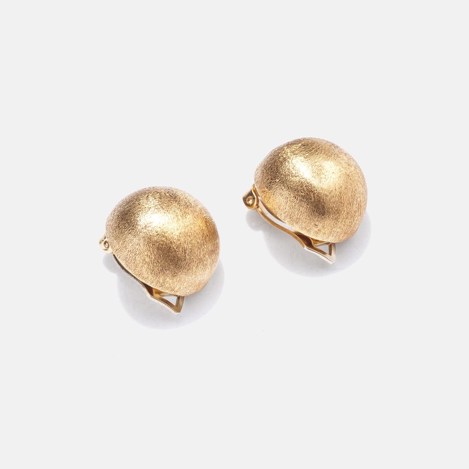 These 18 karat gold clip-on earrings feature a distinctive dome shape, presenting a subtle but appealing volumetric form. Their surface is finished with a matte, brushed texture that softens the gold's natural sheen, adding a contemporary touch to