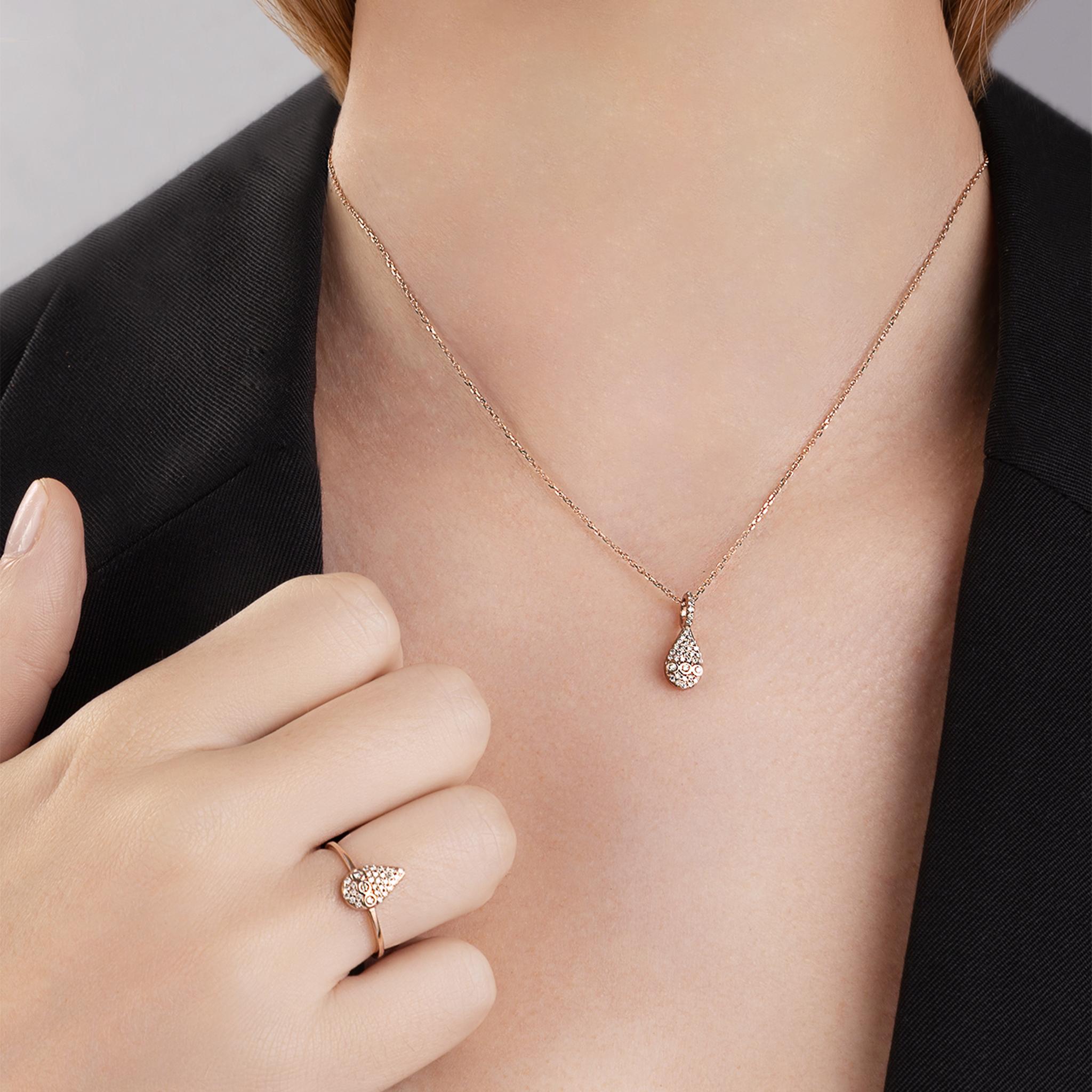 Este impresionante anillo de gota plana en reluciente oro rosa de 18 quilates evoca la maravilla de la naturaleza al brillar como una gota de agua, prístina y pura en el suave abrazo de la mañana. Su disposición de diamantes champán en pavé está