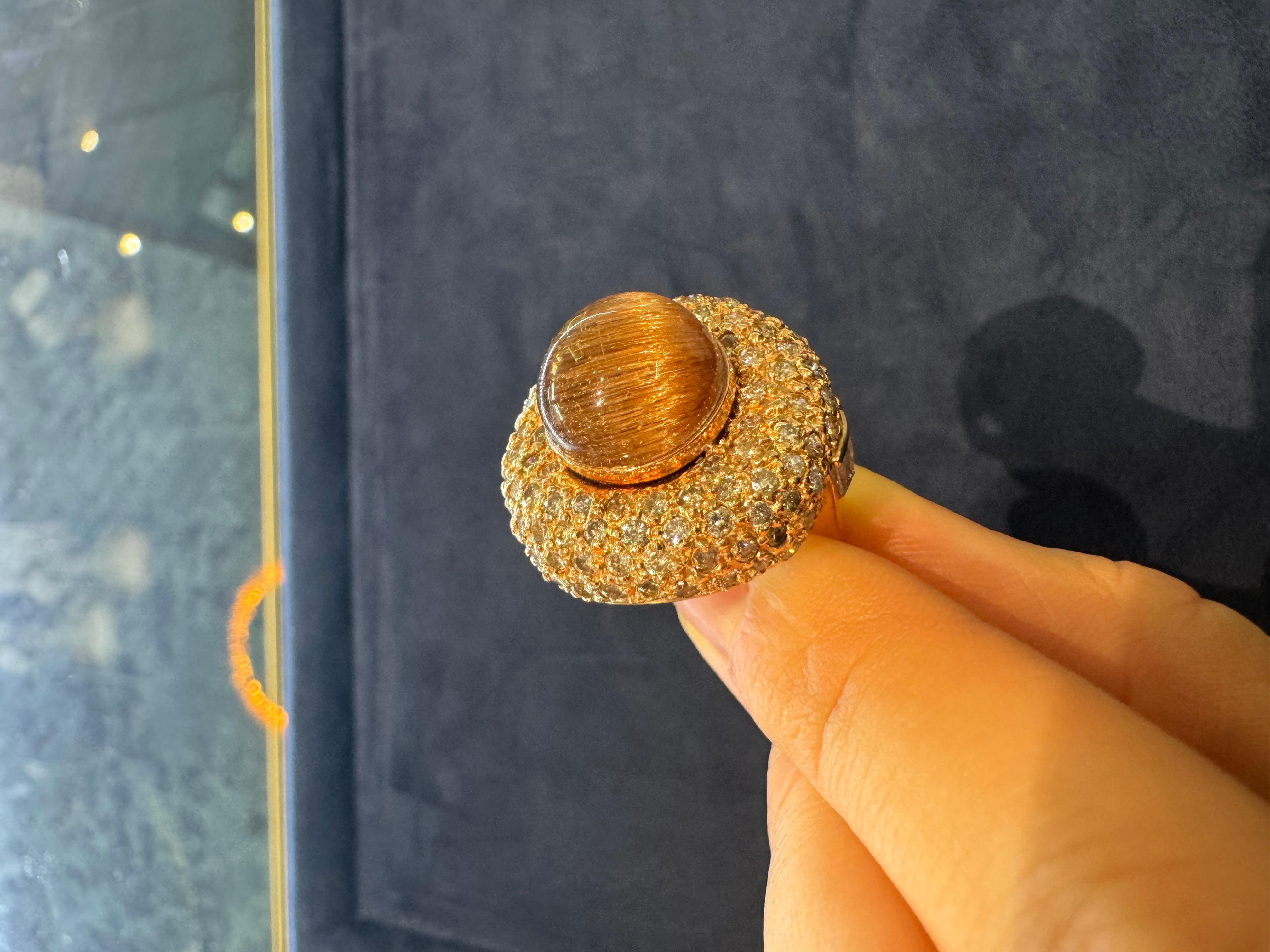 Questo splendido anello in oro rosa 18 carati è un vero e proprio capolavoro, con un sorprendente quarzo rutilato cabochon da 23,49 carati come pietra centrale. I filamenti unici e dorati di rutilo all'interno del quarzo creano un motivo unico nel
