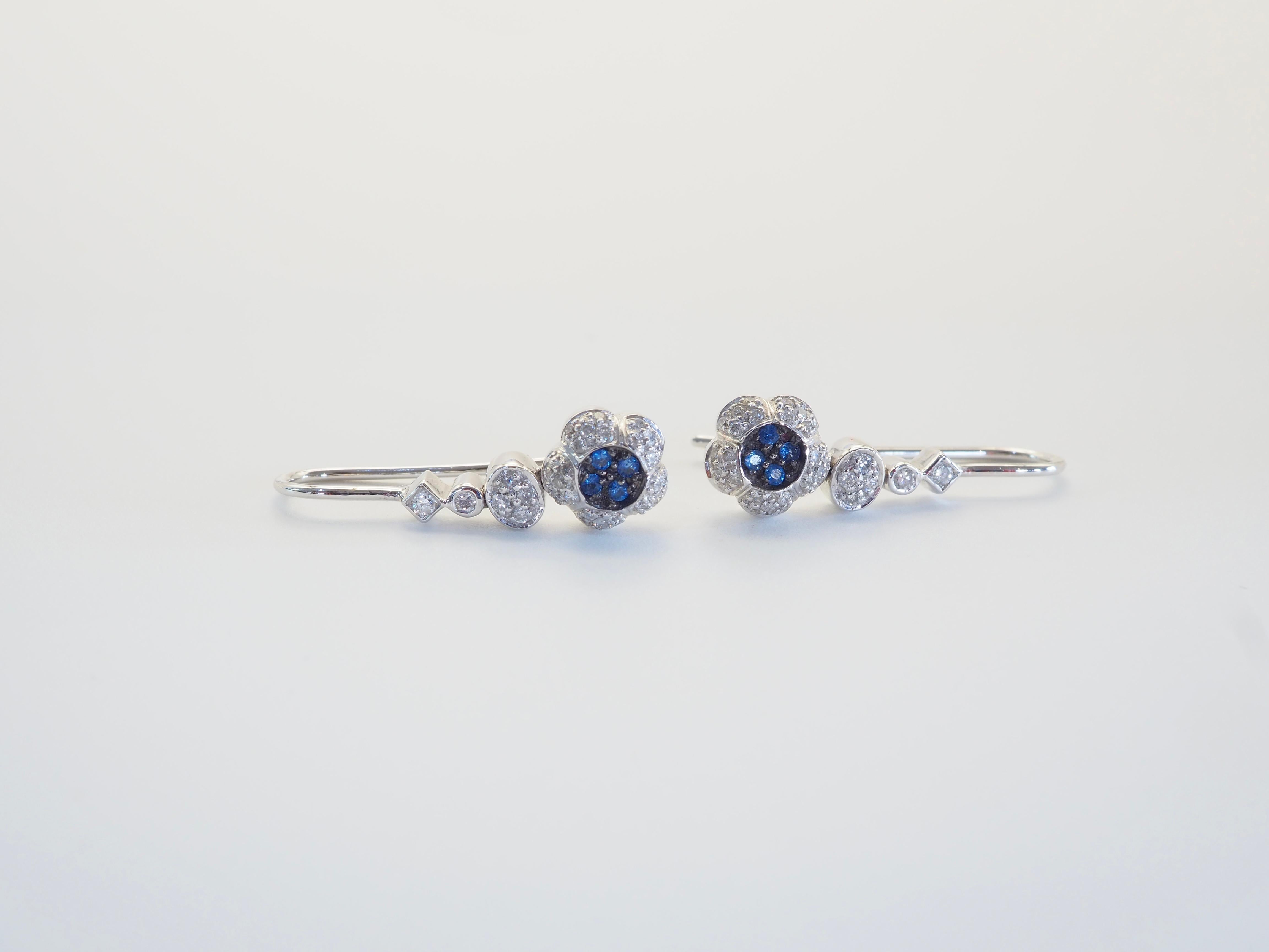 A refined pair of floral dangle earrings crafted in luminous 18K white gold, showcasing the artistry of timeless design. Each earring features a delicate cluster of 0.09 carats of vivid blue sapphires, encircled by a sparkling cluster of 0.31 carats