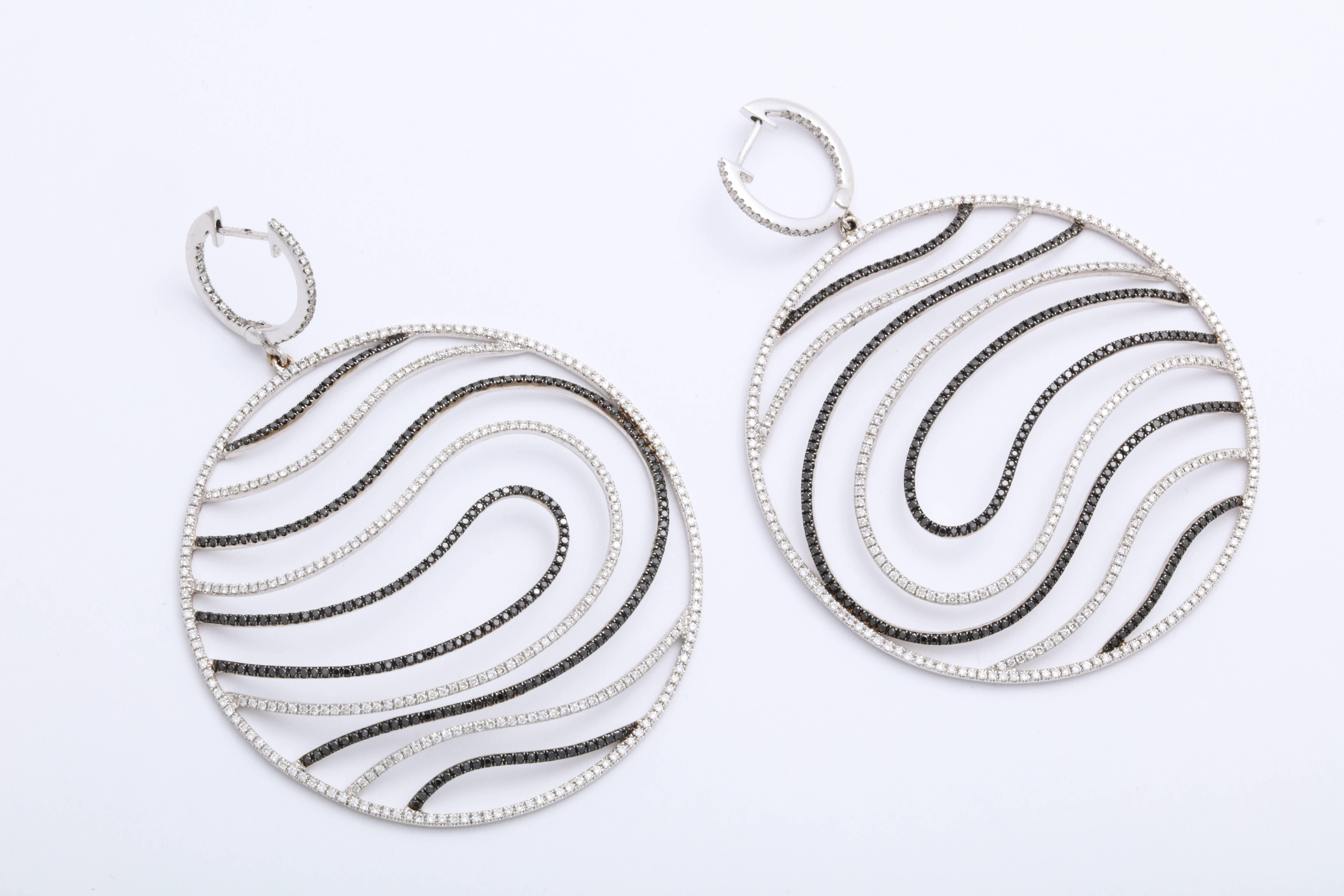 These showstopping earrings are 18 Karat white gold hoops that suspend over-sized circular disk pendants decorated with round black diamonds alternating with colorless diamond free-form waves: 3.58 carats combined total weight.