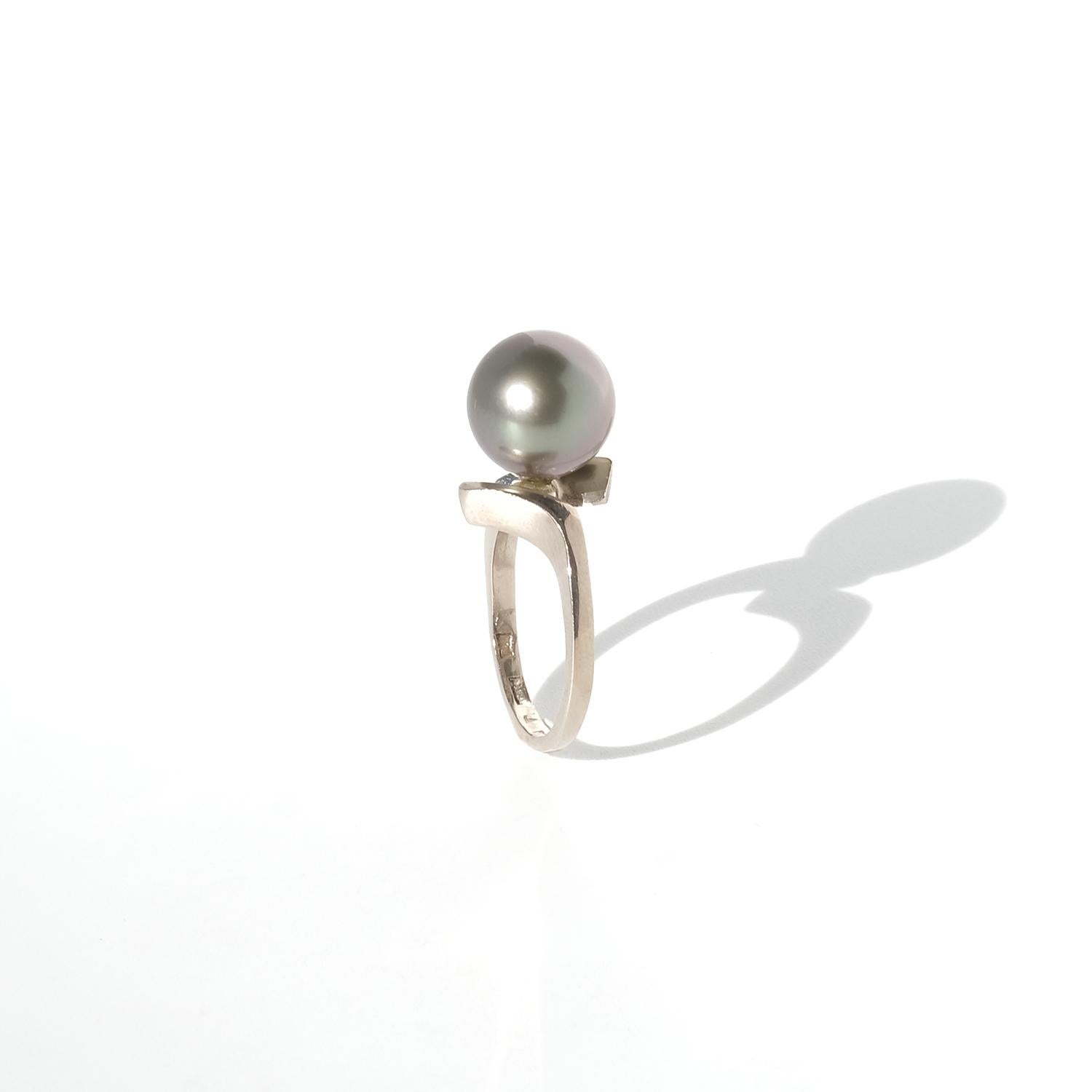 This white gold ring features a lustrous Tahitian pearl with a smooth, perfectly round shape and a rich gray hue that reflects subtle green and silver undertones. The band has a sculptural, asymmetrical design, with a gentle curve and a flat,