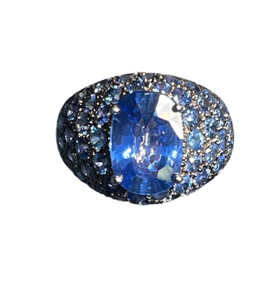 This is an 18K white gold blue sapphires dome cocktail ring. It has an oval cut faceted blue sapphire prong setting in the center, whose measurements are 11.98 x 8.35 x 4.58mm and its weight is approximately 3.78 carat. Pave cluster of fifty one