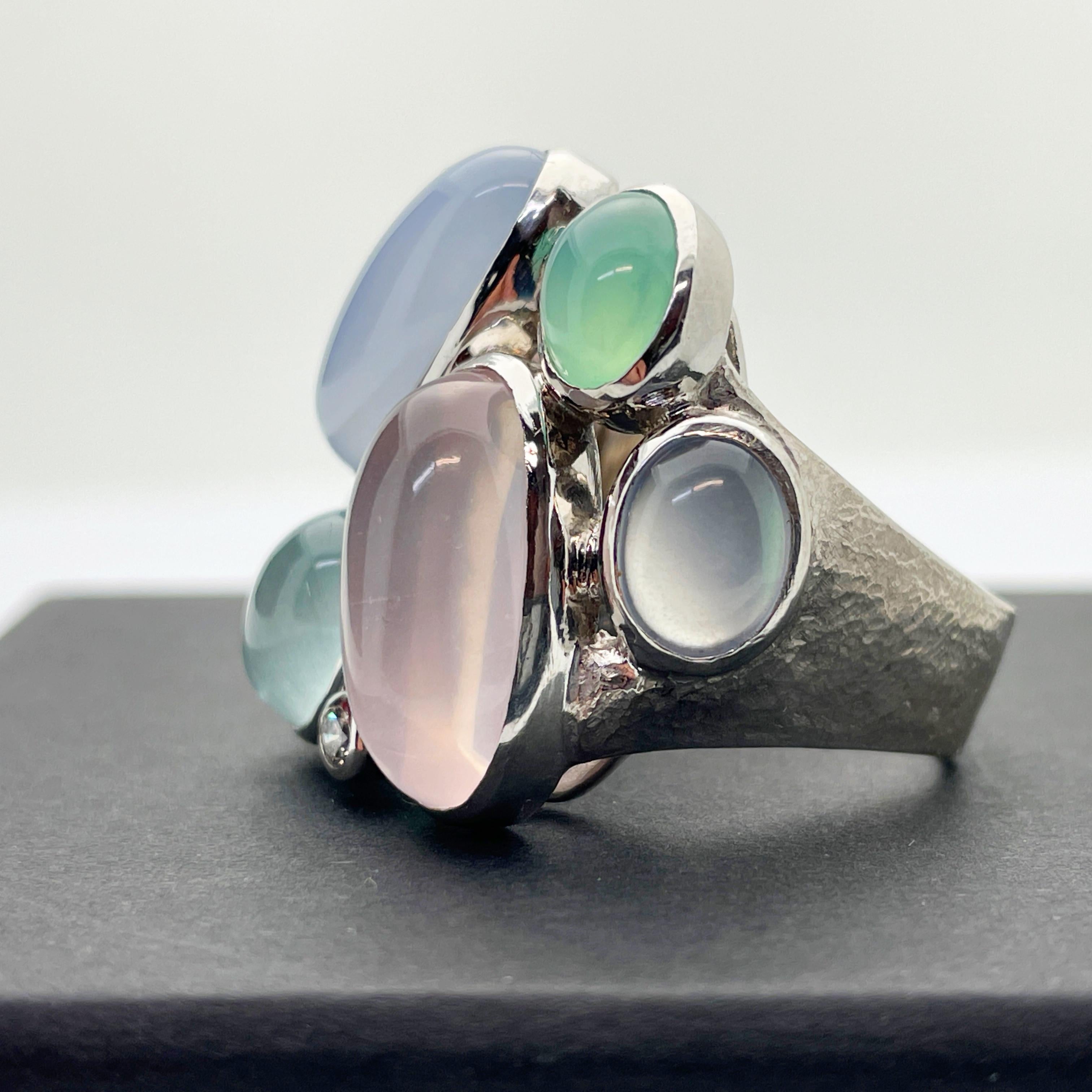 Here is a beautiful and substantial 18k White Gold Custom Chalcedony and Diamond Cluster Ring.

This ring features 7 x oval cabochon cut chalcedony stones ranging in colour from slight violetish blue, green and pink. Also included are 2 x 0.04ct