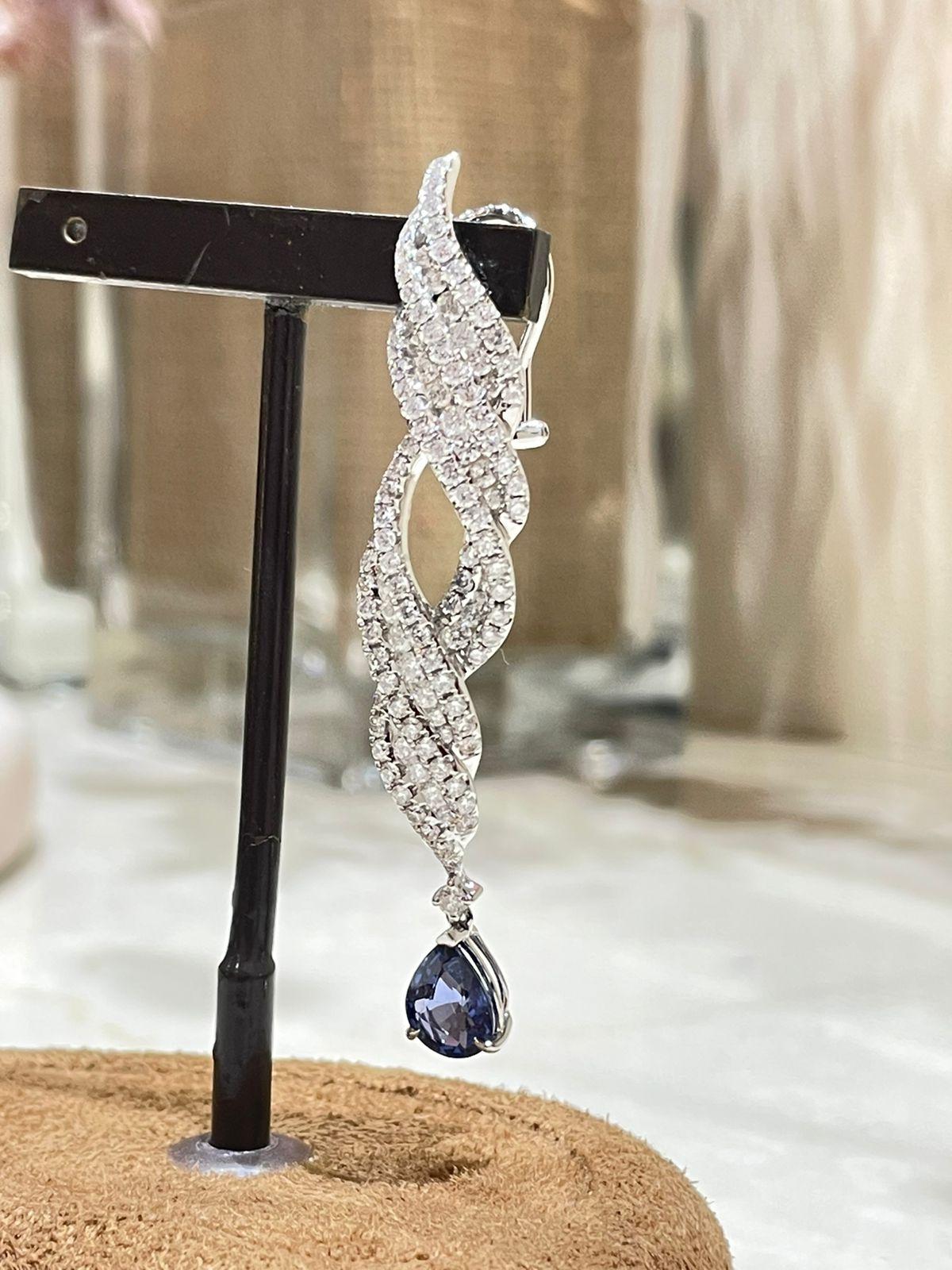 These dangling earrings exude sophistication and elegance with a perfect balance of simplicity and opulence. A striking blue sapphire takes the stage adorned with exquisitely round-cut diamonds. The design ensures a reflection of light and glamour
