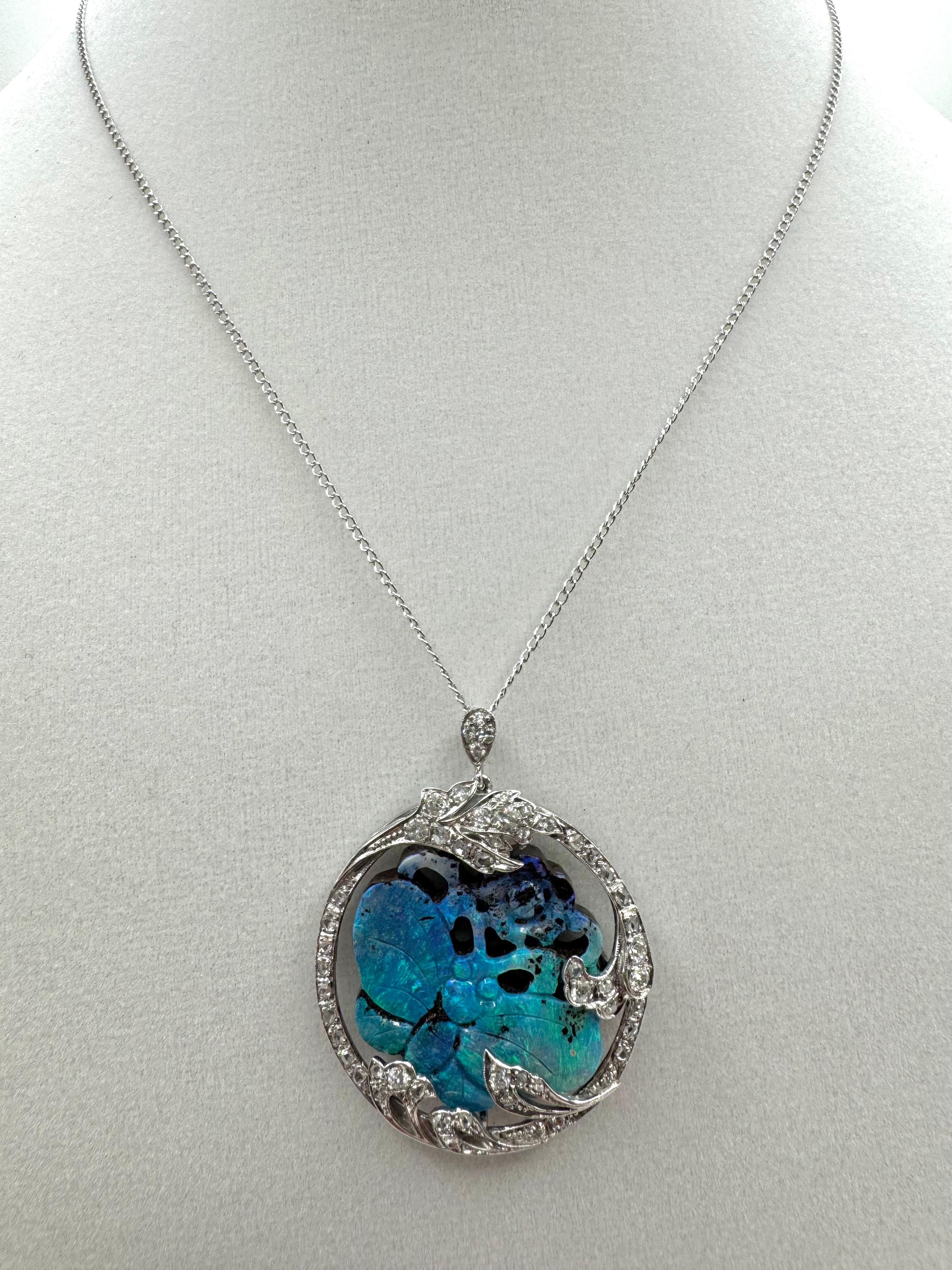 A breathtaking vintage necklace featuring a meticulously carved butterfly from natural opal, glowing with vivid blue and green hues. The opal centerpiece is surrounded by an ornate, nature-inspired vine frame set in rich 18K white gold and adorned