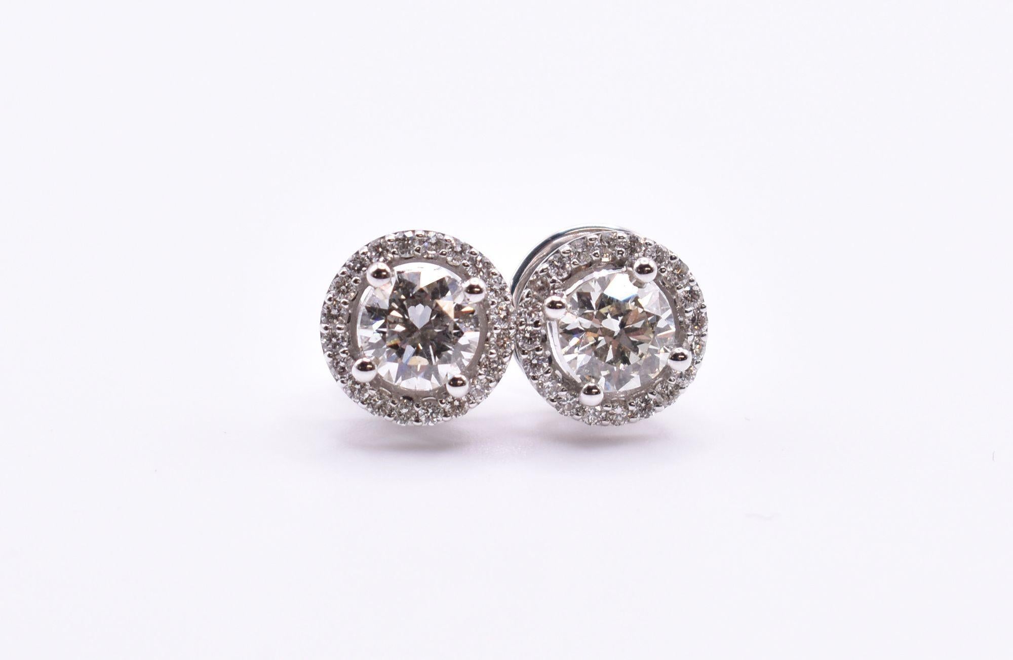 A splendid pair of 18 karat white gold diamond halo stud earrings featuring two bezel set round brilliant cut diamonds of SI1 clarity clarity and I/J colour totaling 1.18 carats, surrounded by 18 prong set diamonds totaling an additional 0.25