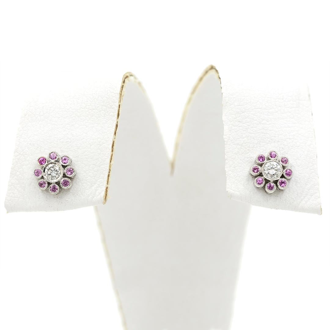 This pair of 18 karat white gold stud earrings feature a round brilliant cut diamond at their center, weighing a combined 0.12 carats, surrounded by eight round cut pink sapphires, weighing a combined 0.16 carats. The earrings have posts and
