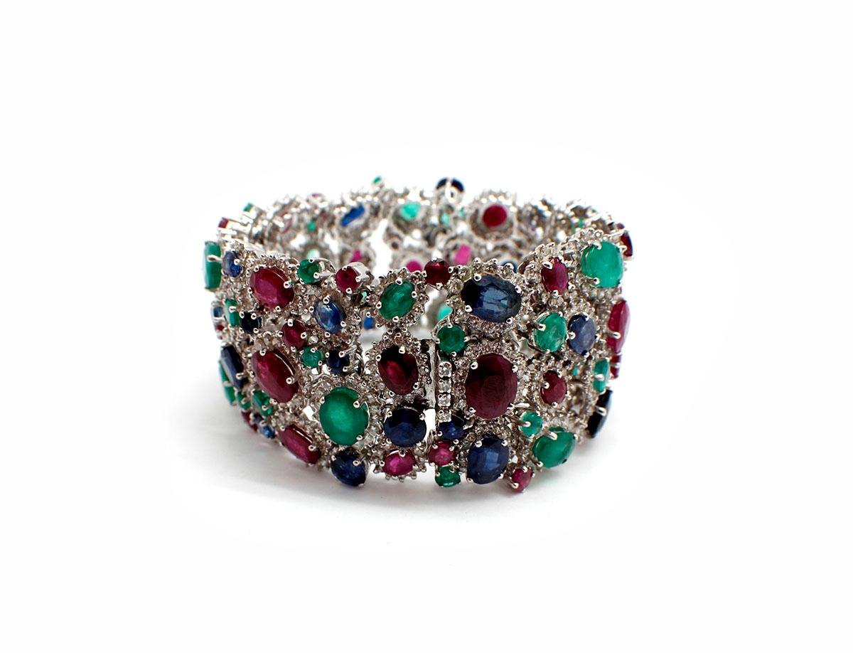 This stunning bracelet is made in 18k white gold. It holds various sizes of oval-cut rubies, emeralds, and sapphires. All the gemstones are accented by smaller round brilliant diamonds. The gemstones have a total weight of 83 carats, and the