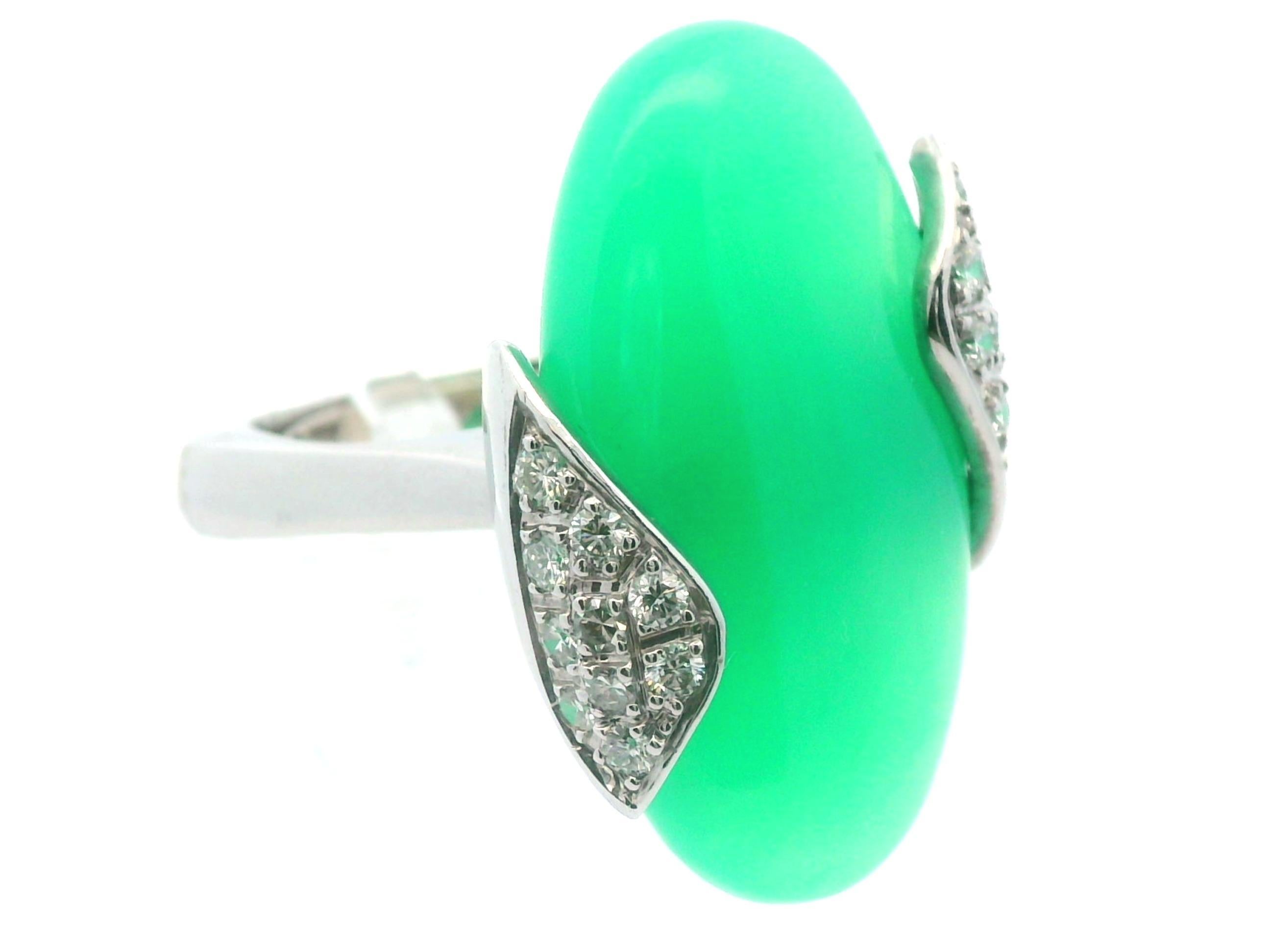 Este impresionante anillo de crisoprasa y diamantes se fabricó en la década de 1950 en oro blanco de 18 quilates. La piedra central es una vibrante crisoprasa ovalada de 24 quilates que cautiva la atención. Tanto el lado izquierdo como el derecho