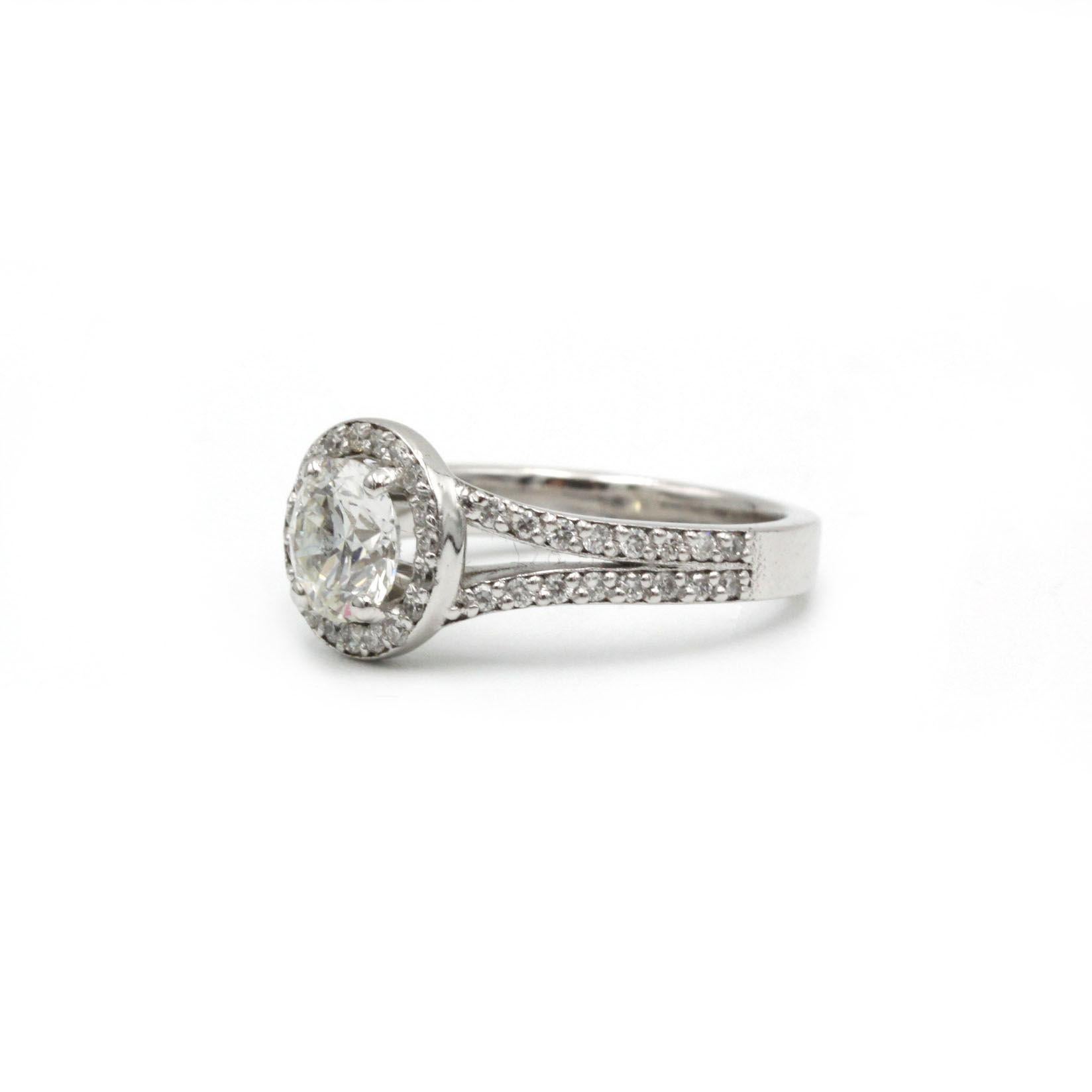 Espressione di un'eleganza senza tempo, questo anello solitario in oro bianco 18 carati è incentrato su un diamante rotondo brillante da 0,99 ct H VS1, il cui fuoco è amplificato da una lucente aureola di pietre più piccole dal taglio brillante. La