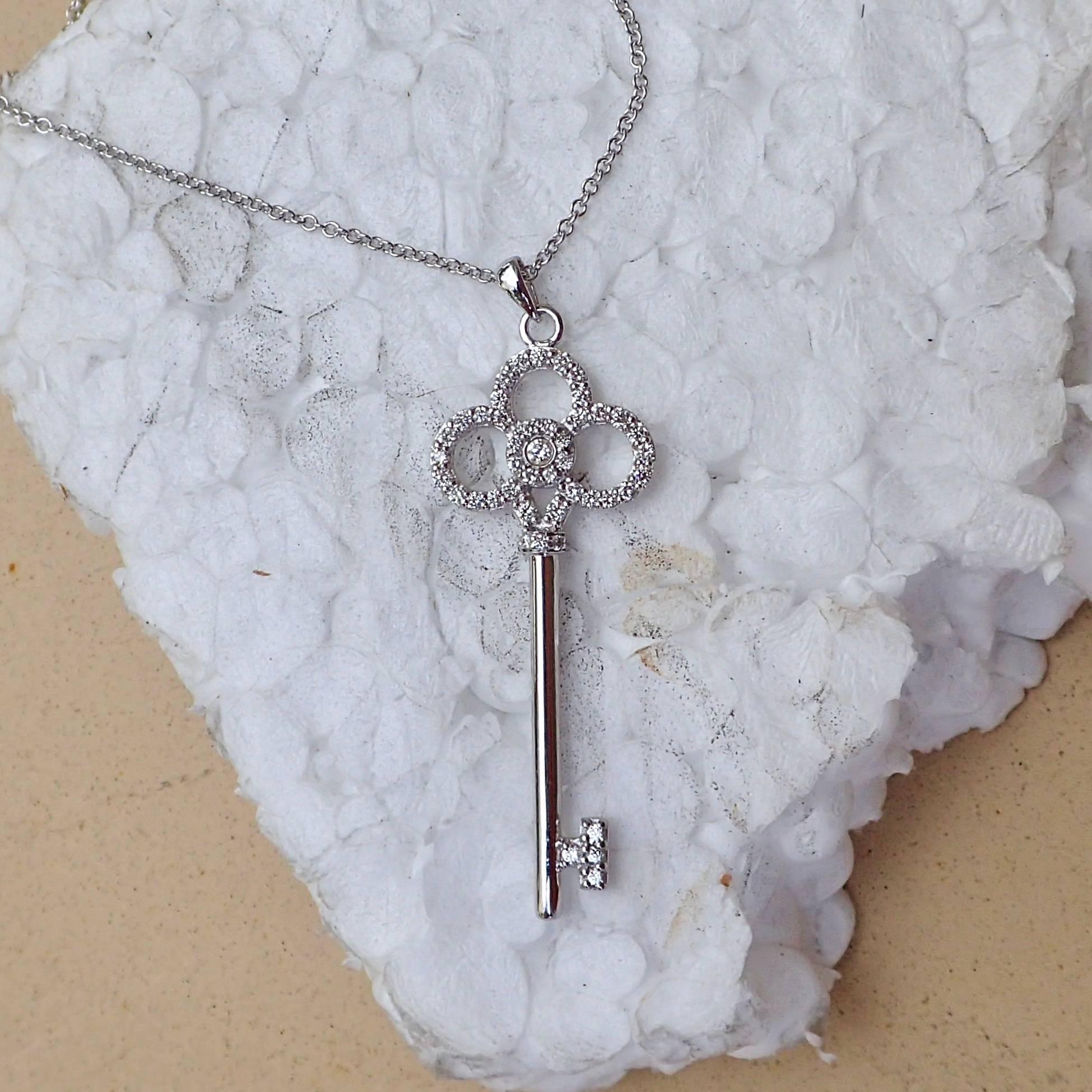 18k White Gold Key shaped pendant hangs from an 18k white gold 1.5mm cable chain. The pendant is set with one (1) 1.6mm Round Brilliant Cut diamond weighing 0.02 carats with Clarity Grade VS and Color Grade E-F and twenty-one (21) 1.3mm Round