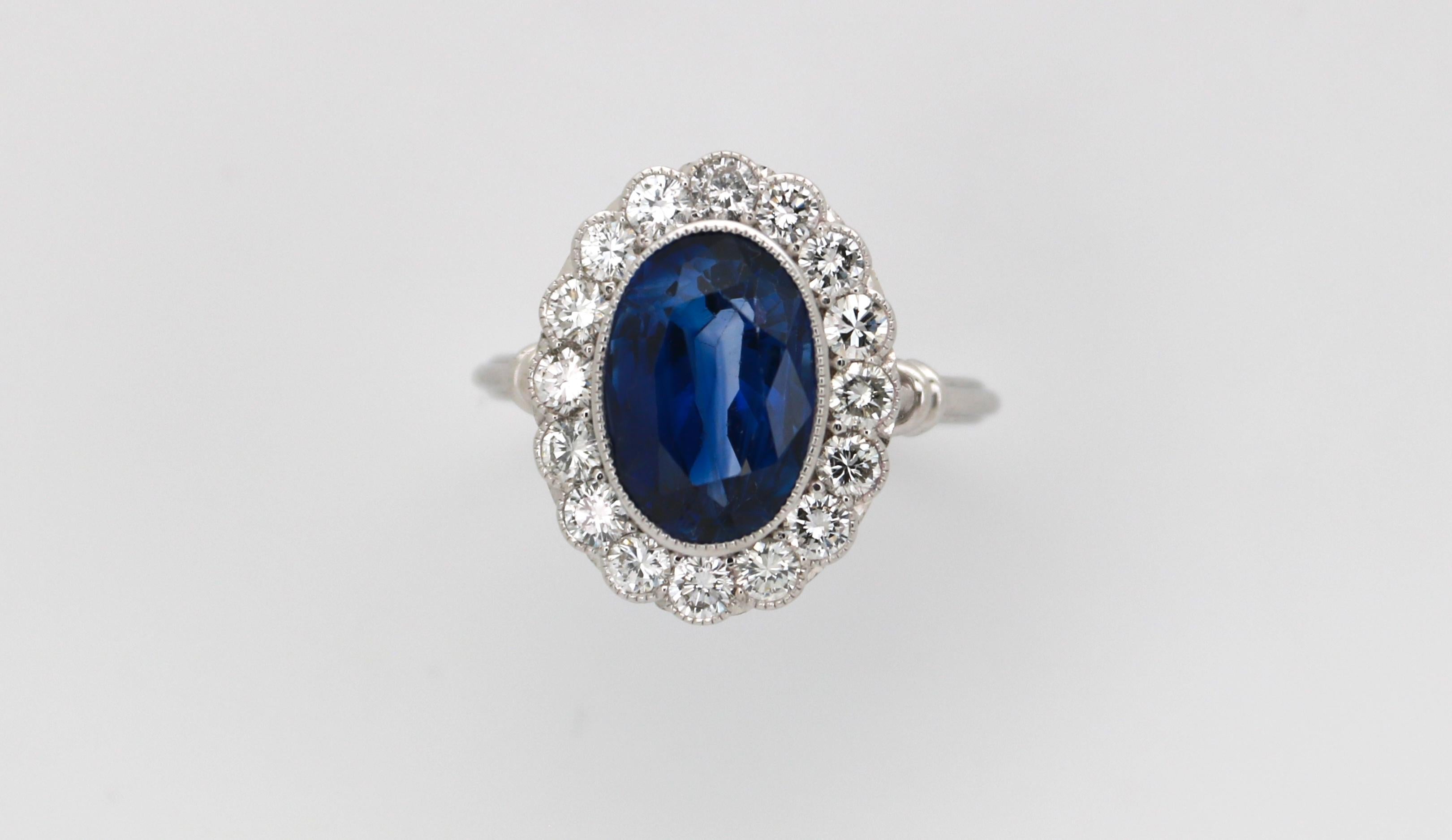 Un anello a grappolo / entourage in oro bianco 18k, unico e fatto a mano, con al centro uno zaffiro blu vivo naturale di taglio ovale del peso di 6,16 carati e circondato da 16 diamanti taglio brillante per un peso totale stimato di 0,80 carati. Lo