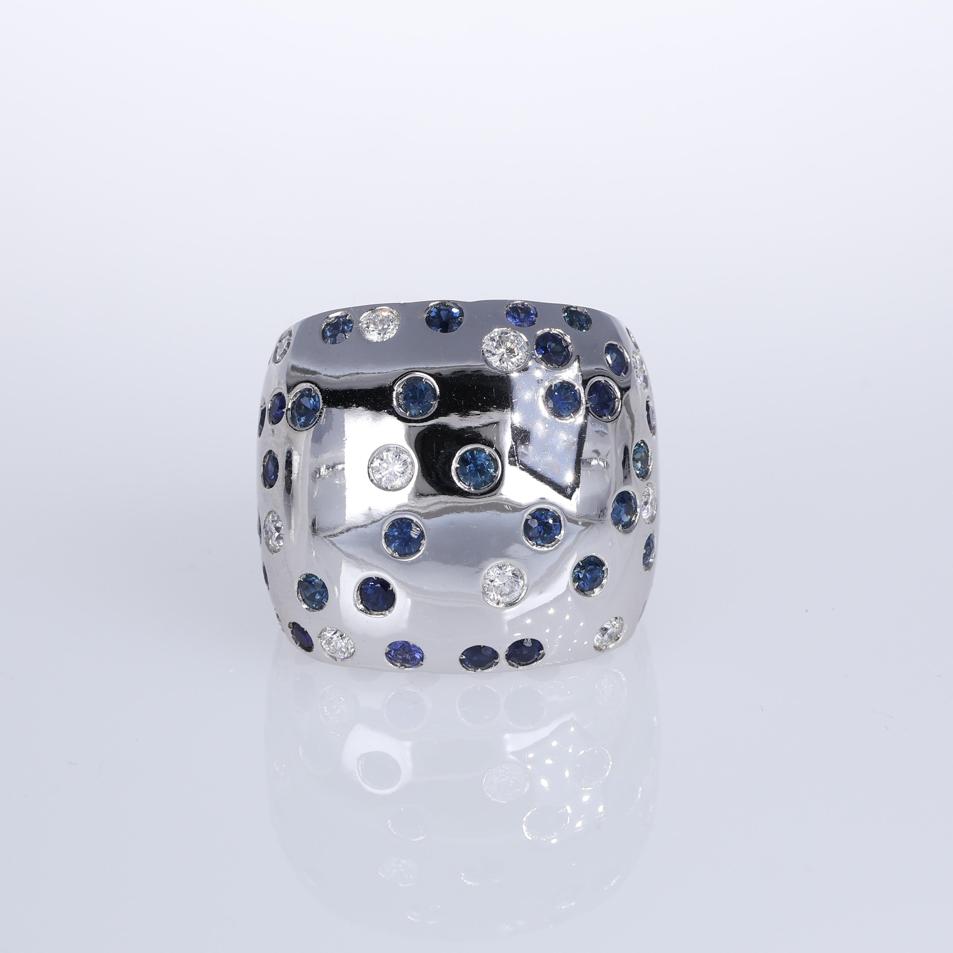 A modern and artistic creation, this 18K white gold ring features a playful polka dot pattern of vibrant blue sapphires and brilliant-cut diamonds. The polished gold surface provides a sleek backdrop, highlighting the contrast and brilliance of the