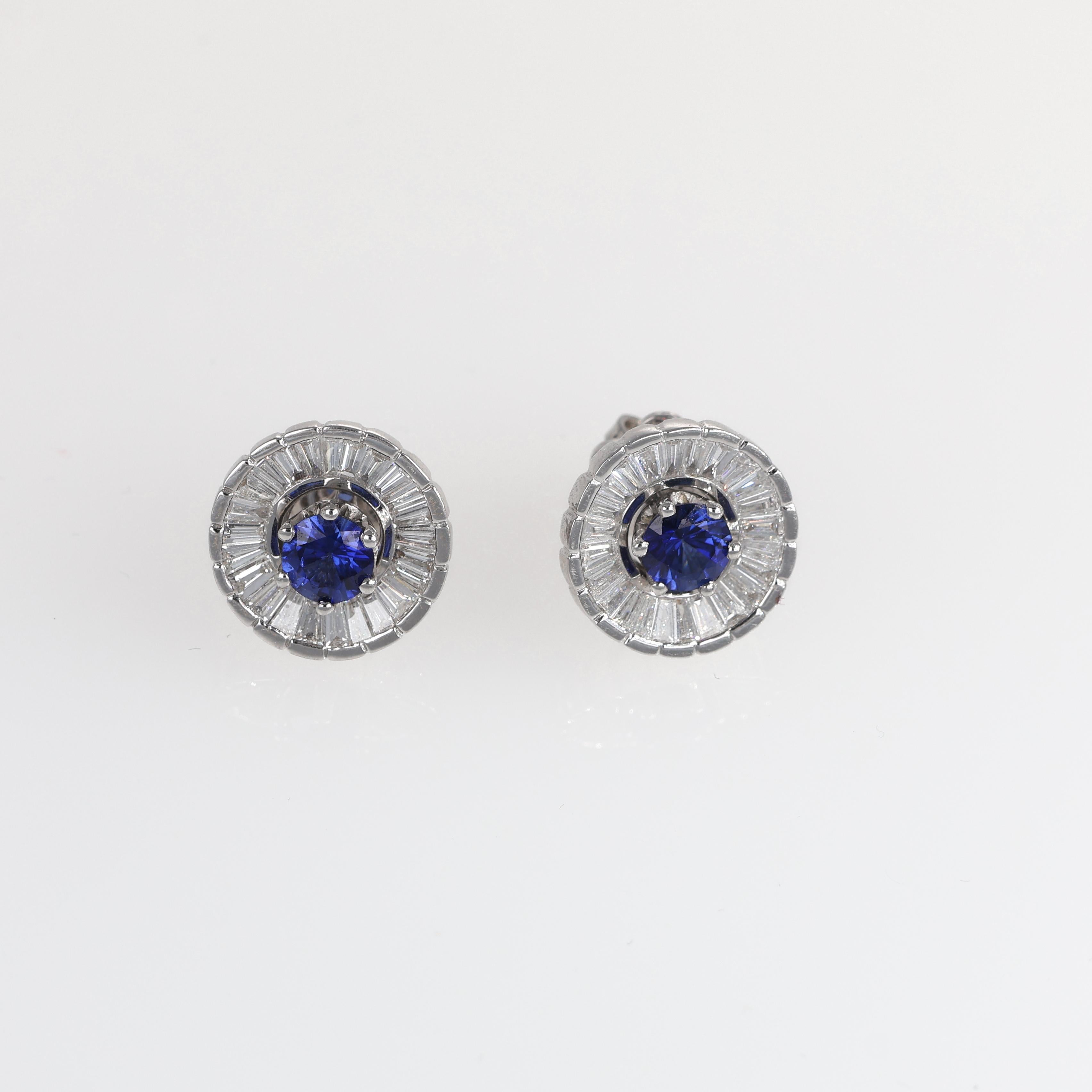 A timeless pair of stud earrings crafted in 18K white gold, featuring a central round-cut natural blue sapphire, elegantly surrounded by a halo of baguette-cut white diamonds in a spinning pinwheel design. The contrast between the deep blue