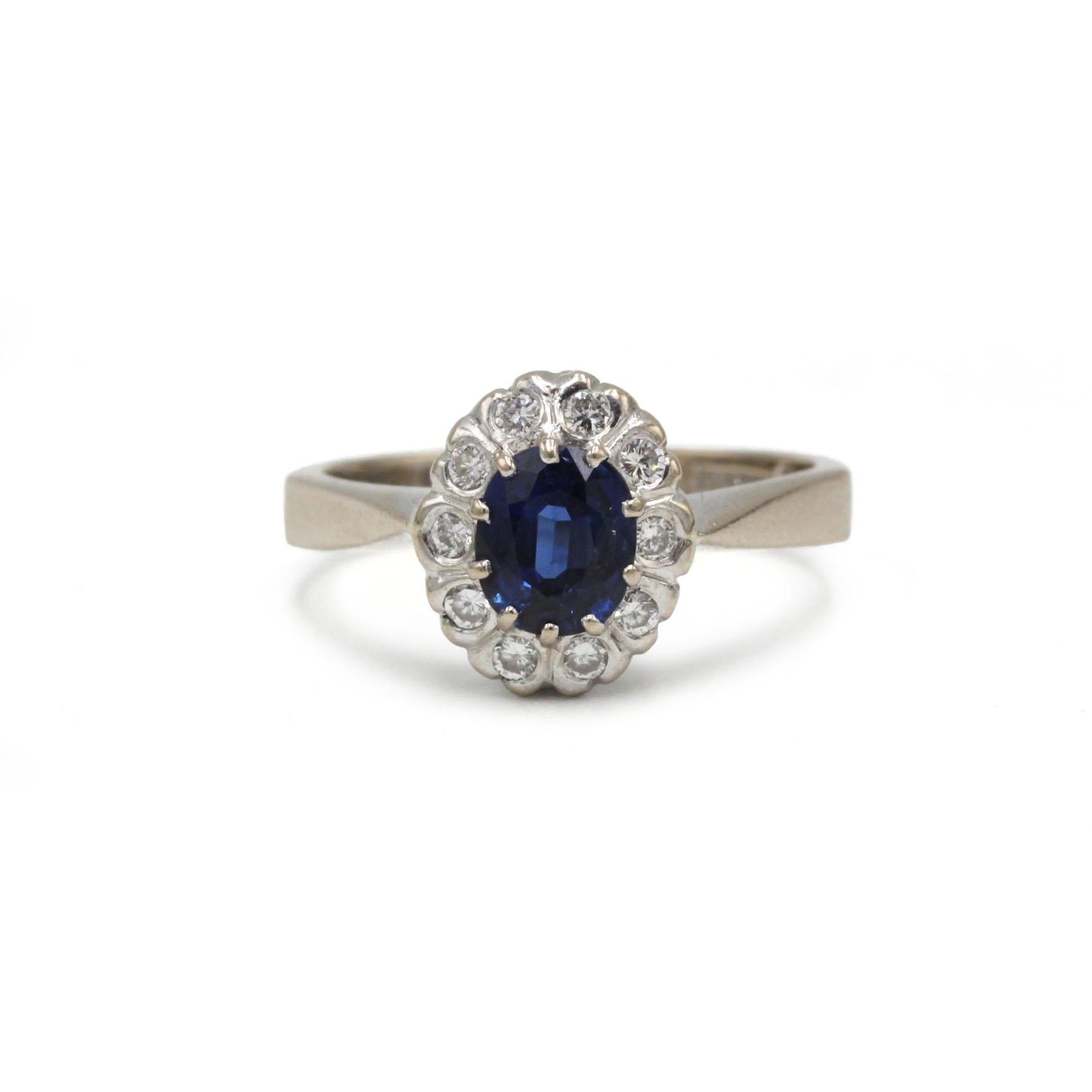 Dieser elegante Ring aus 18-karätigem Weißgold zeigt in der Mitte einen lebhaften, natürlichen blauen Saphir von 0,93 Karat, der von einer Gruppe runder Brillanten eingerahmt wird. Das zeitlose, ovale Cluster-Design hebt die Farbtiefe des Saphirs