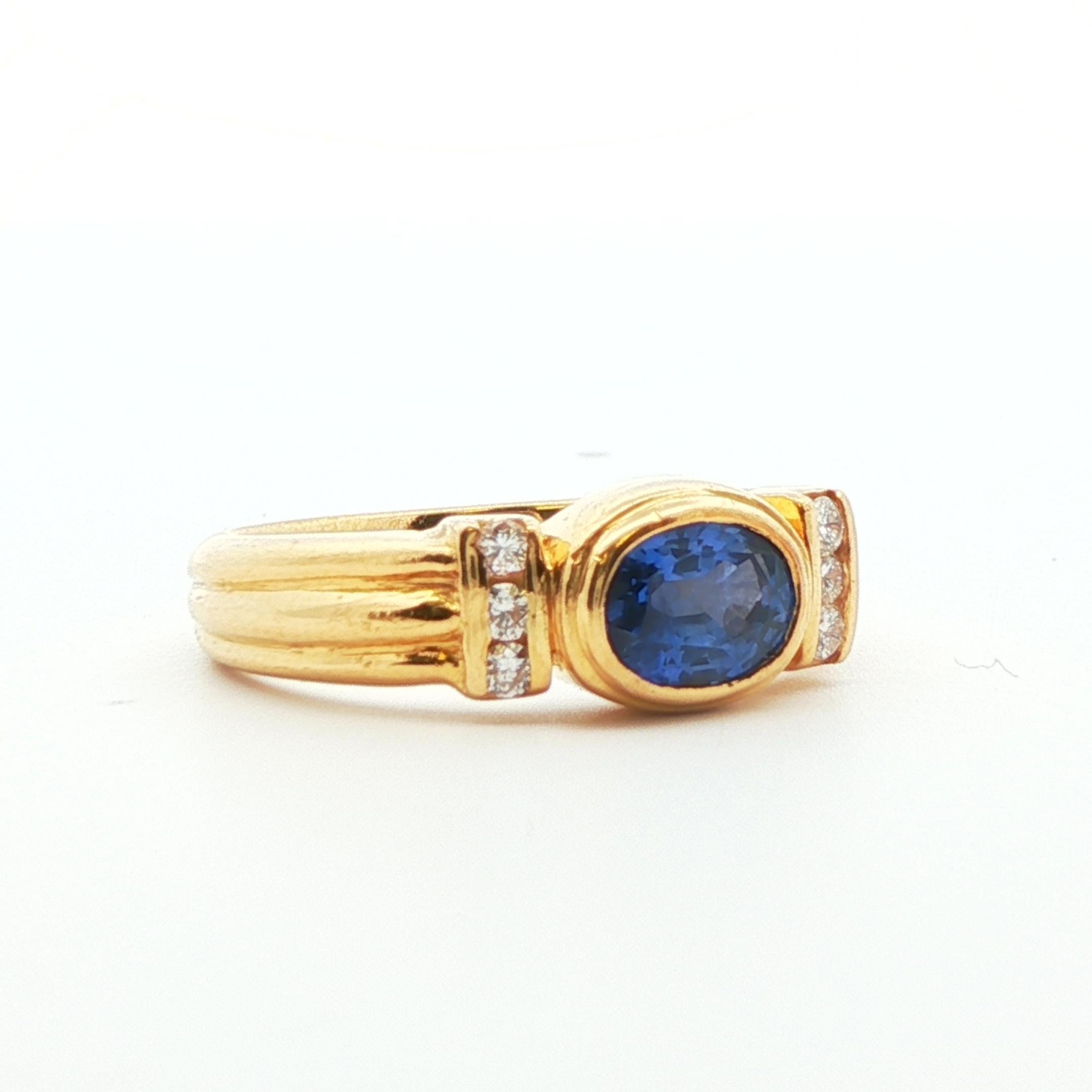 Este impresionante anillo está hecho en oro amarillo de 18 quilates con zafiro azul natural y diamante. El centro del anillo exhibe un zafiro azul intenso de talla oval engastado en un bisel con una fila de 3 diamantes engastados en cada lado, lo