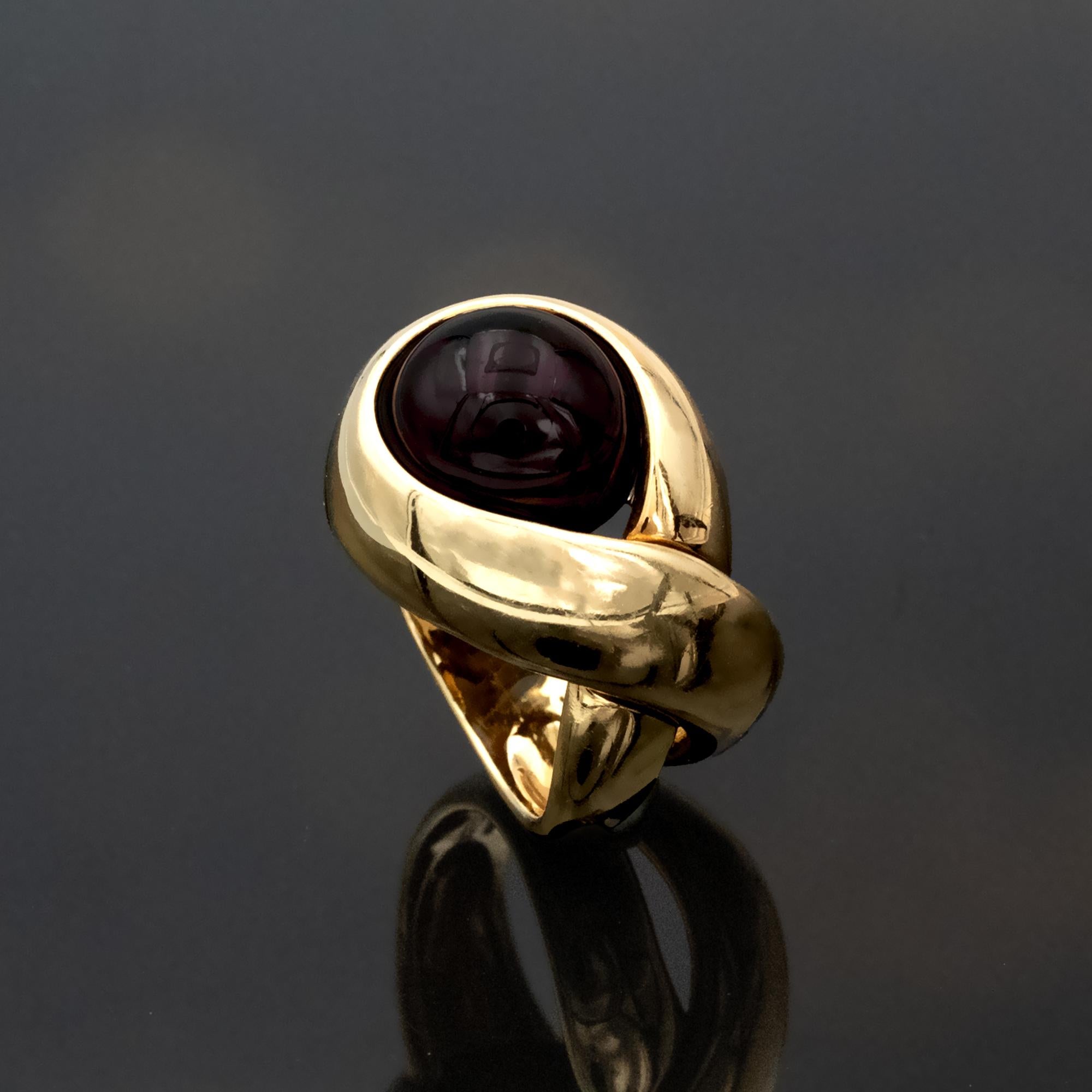 This ring, offered by Claris-A, in solid yellow gold features a rounded cabochon garnet set in a protective bezel that keeps the stone fully visible while securing its edges. The gold structure wraps around the garnet in an asymmetric, partially