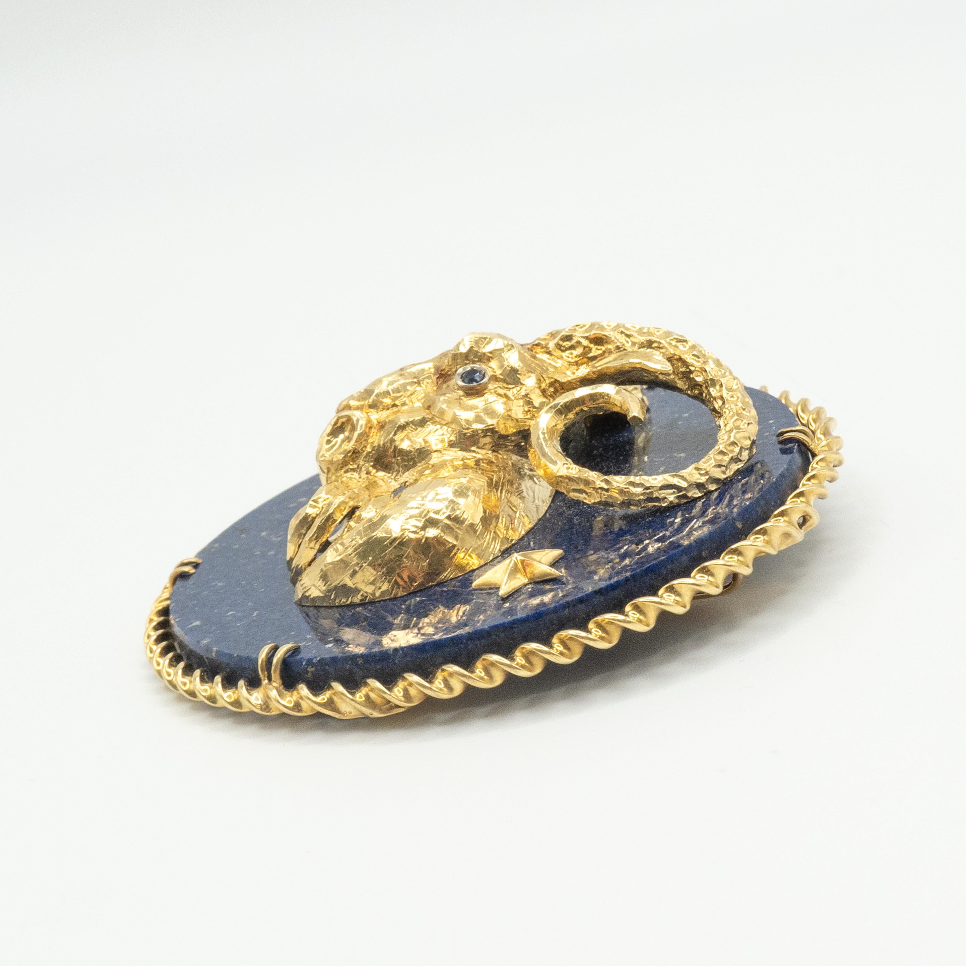 This is a rare and collectible 18K gold Capricorn zodiac brooch featuring a richly textured, three-dimensional goat’s head with curling horns, mounted on a deep blue lapis lazuli disc with gold star accents. The eyes set with round facet sapphires,