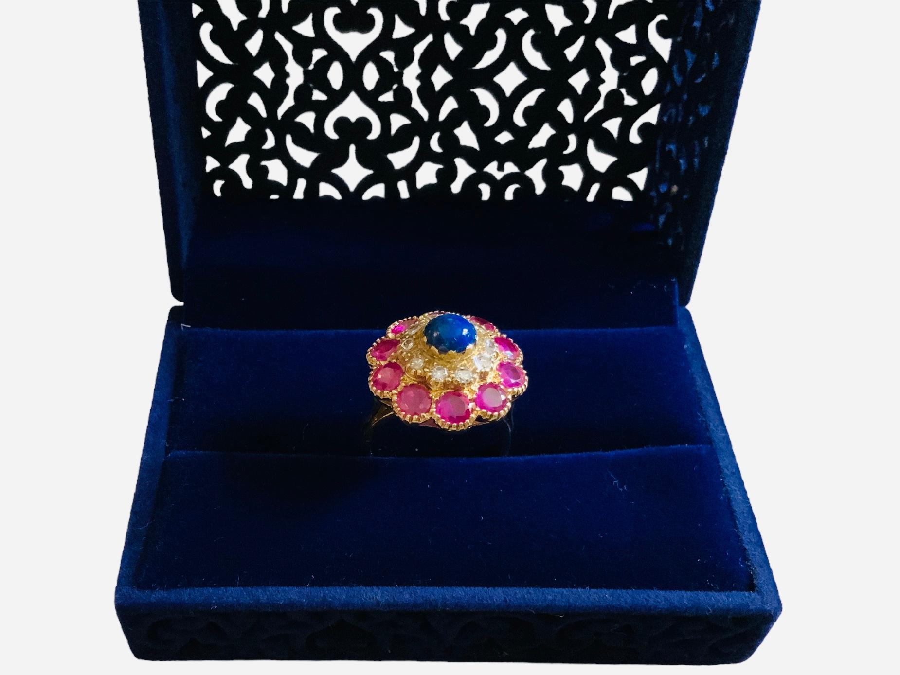 This is an 18K yellow gold diamonds, Lapis lazuli and rubies cocktail ring. The top of the ring forms a flower. This one is made of an external large halo of ten round cut and faceted rubies in prong setting followed by a small halo of ten round cut
