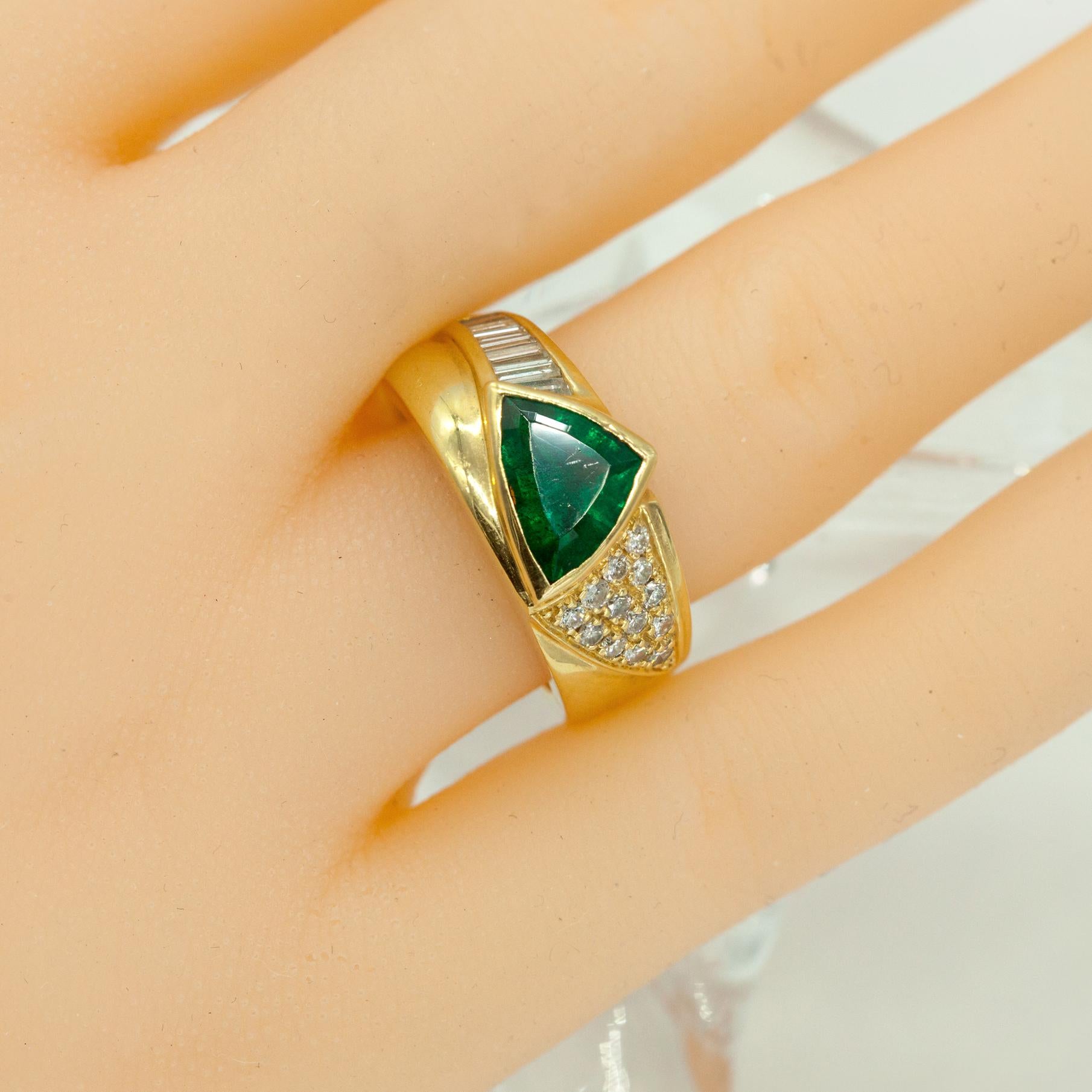 Crafted in rich 18K yellow gold, this bold ring showcases a striking trillion-cut emerald weighing approximately 2.00 carats, with a vivid green hue and natural inclusions typical of emeralds. One side of the emerald is accented by a channel-set row