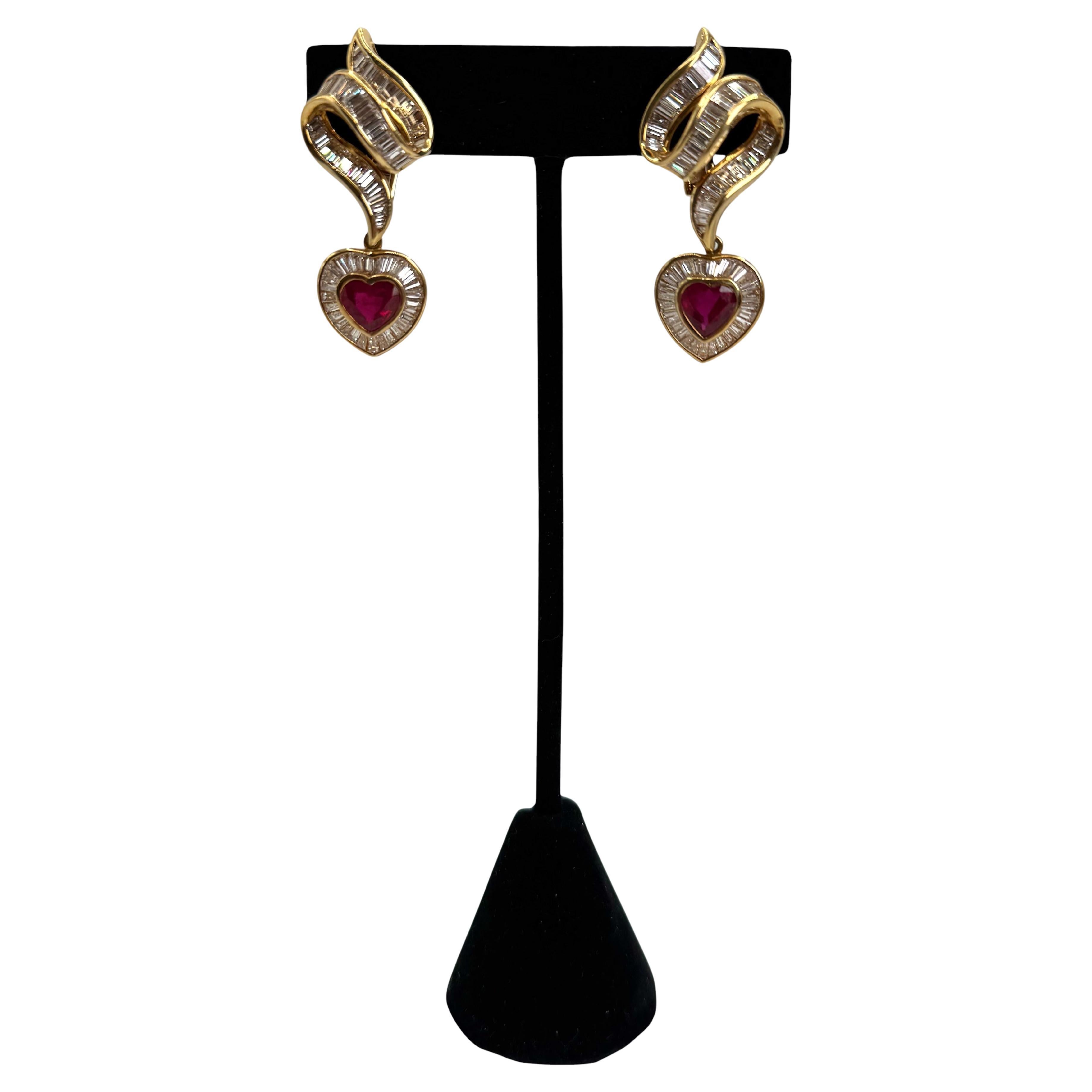 A dazzling celebration of bold romance and refined craftsmanship, these 18K yellow gold earrings feature two richly saturated, heart-shaped rubies totaling approximately 3.49 carats. Each ruby is framed by a radiant halo of baguette-cut diamonds and