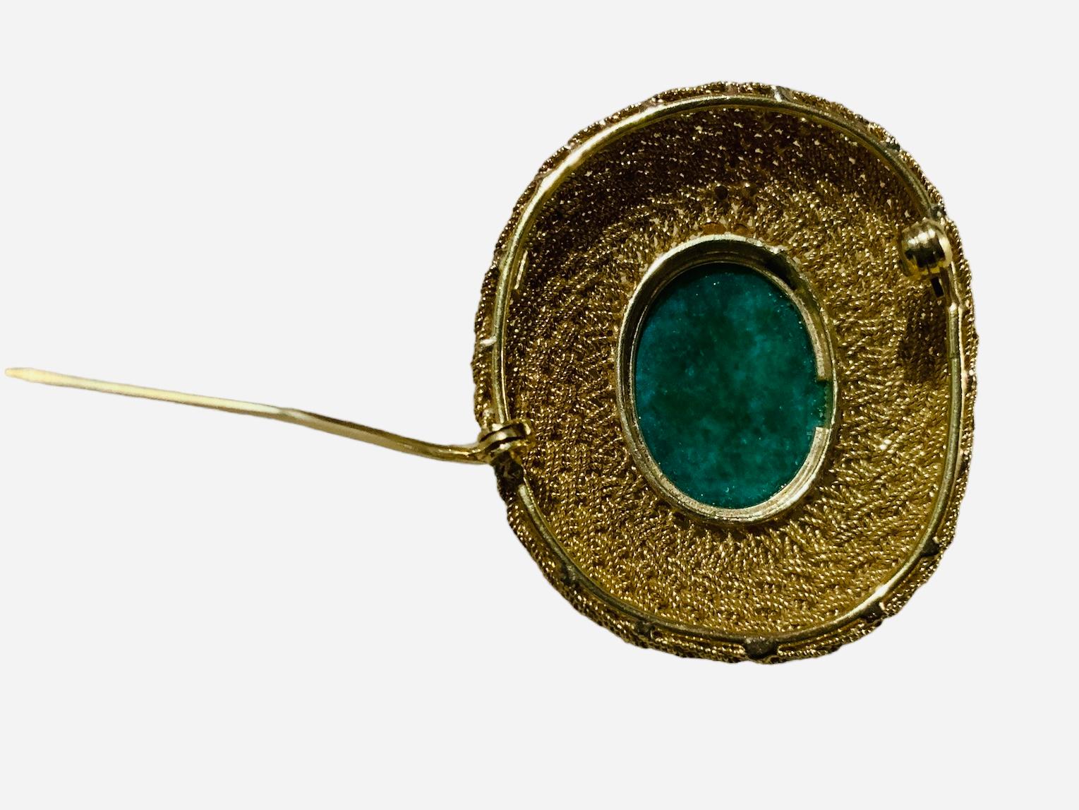 This is an 18K yellow gold Jade brooch. It depicts an oval shaped cabochon jade mounted in bezel setting gold brooch and adorned with gold braided thin ropes. It closes with a C-clasp in its back. It is hallmarked 18k gold in the back.