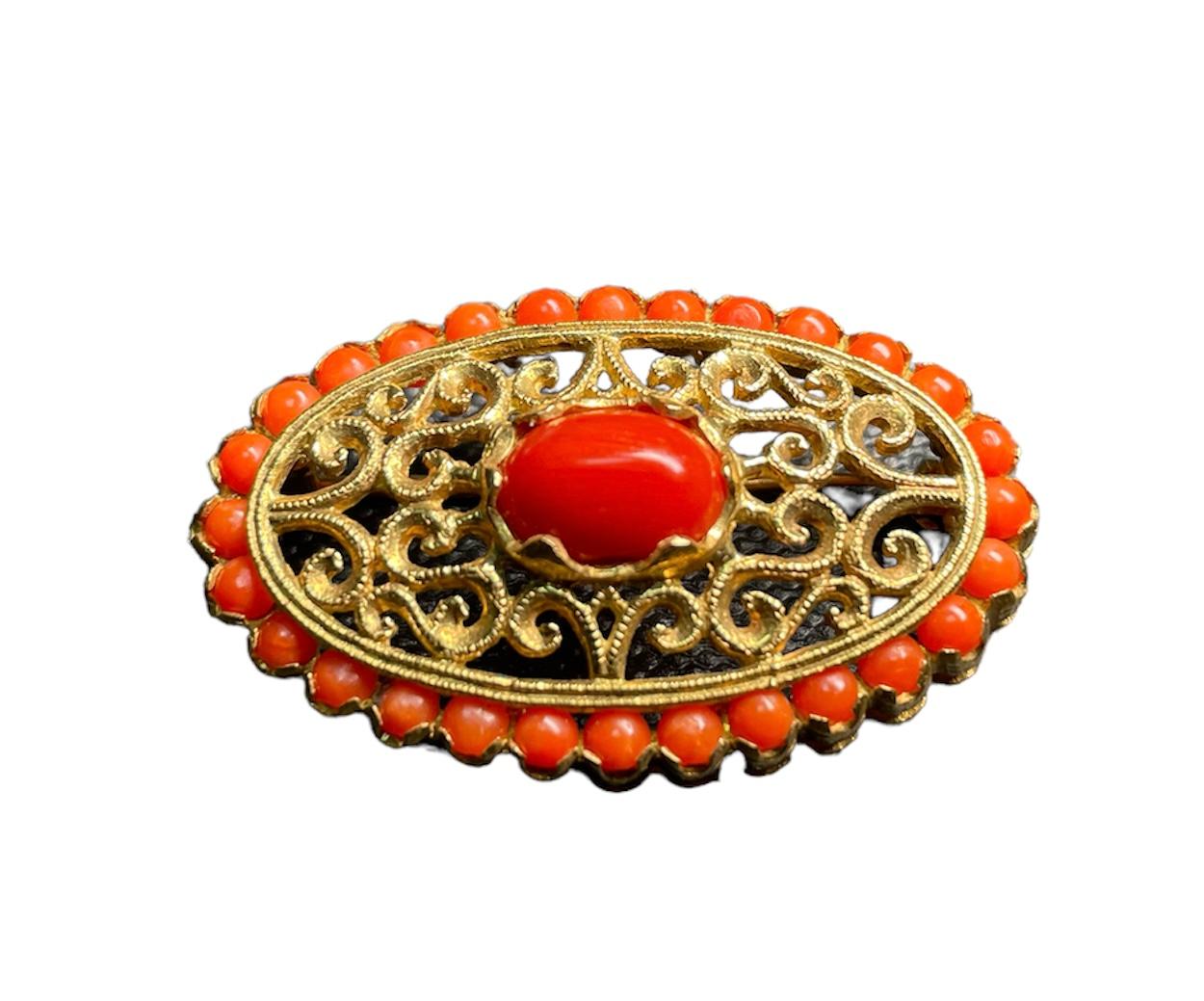 This is an 18k yellow gold and Mediterranean coral brooch. It depicts an elongated oval brooch with a cabochon oval coral in the center in prong setting. Around it, the brooch is adorned with filigree like scrolls. Coral beads over a gold base