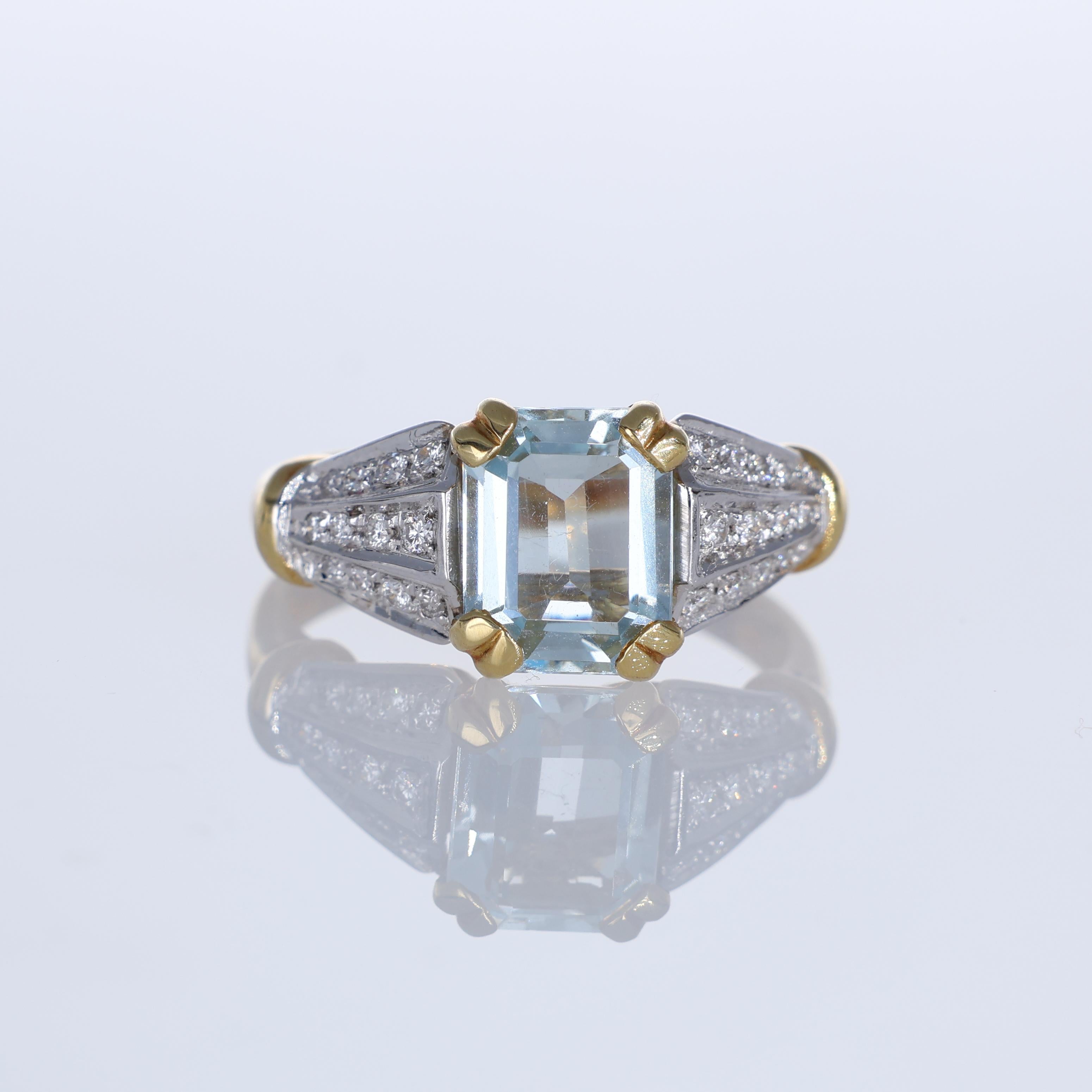 An elegant 18K yellow gold ring featuring an emerald-cut aquamarine, securely held by four yellow gold prongs. The structured design is enhanced by a split shank with diamond pavé, adding brilliance and contrast to the cool blue of the central