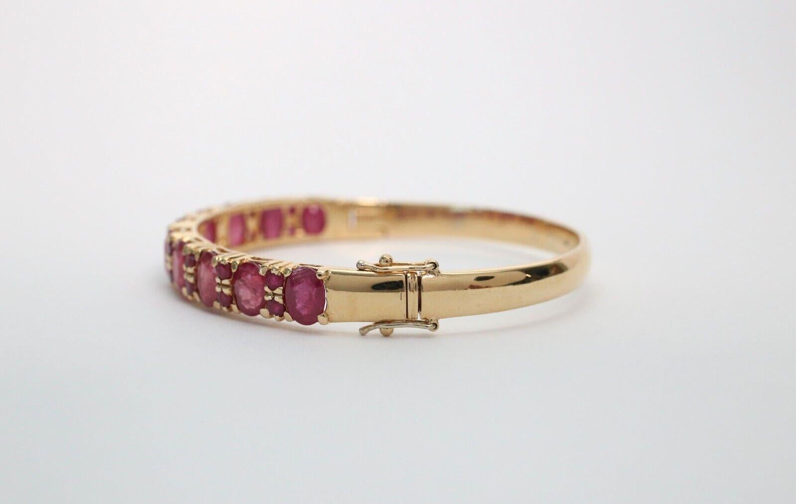 Crafted in rich 18k yellow gold, this elegant bangle showcases a vibrant row of deep pink to red oval-cut stones, prong-set in a classic, symmetrical design. The gemstones are framed by delicate gold separators, adding refined detail and enhancing