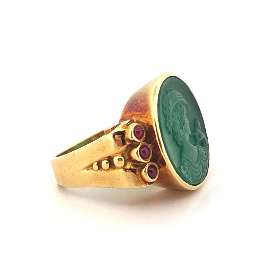 Questo anello in oro giallo 18 carati è caratterizzato da un cammeo in agata verde finemente intagliato che raffigura un profilo femminile classico. L'agata riccamente saturata è accentuata da sei rubini cabochon - tre incastonati su ogni spalla -