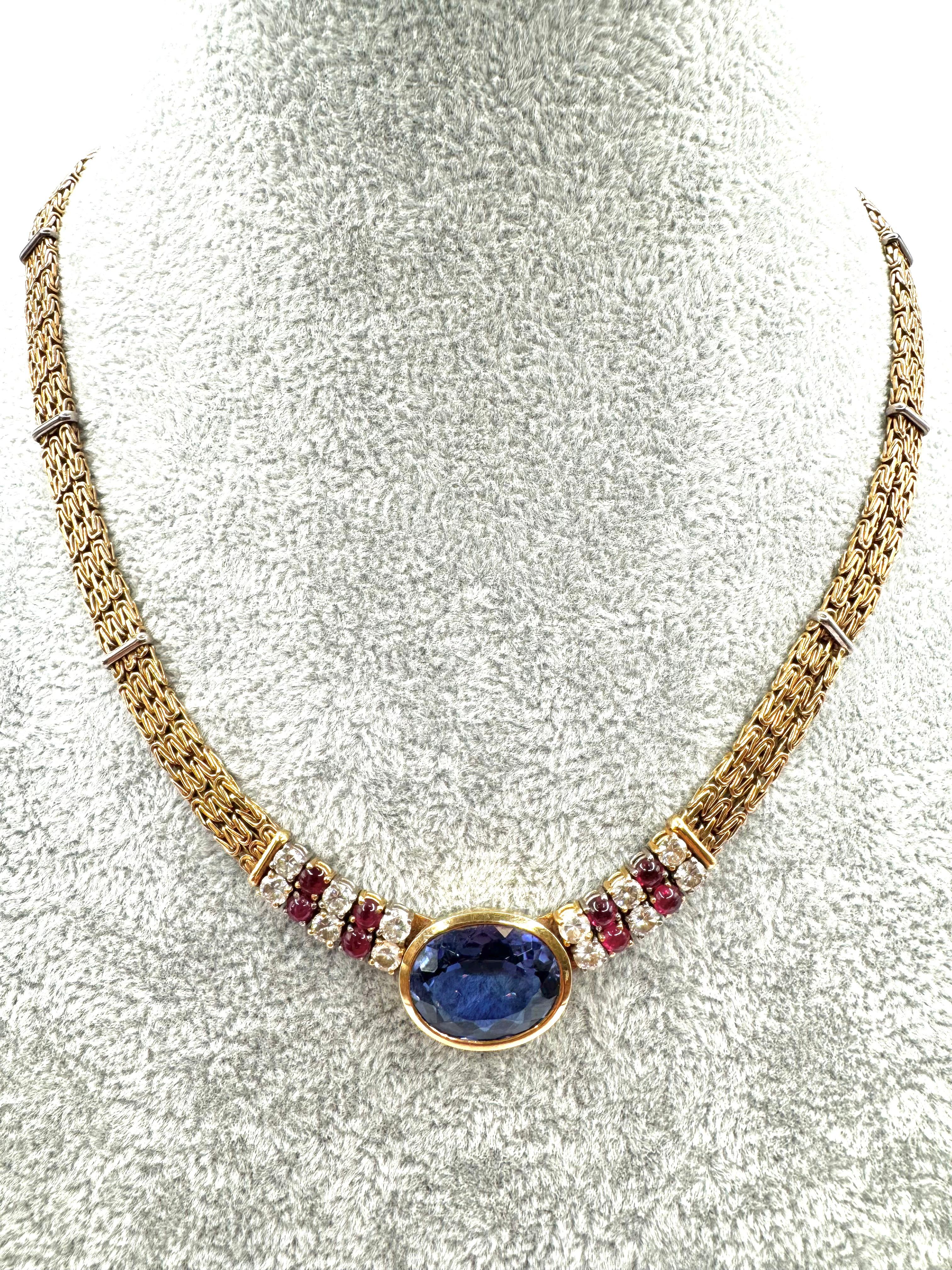 This bold and luxurious vintage necklace, crafted in 18K yellow gold, showcases a stunning oval-cut tanzanite centerpiece with rich violet-blue hues. The collar-style design is flanked by rows of brilliant-cut diamonds and deep red cabochon rubies,