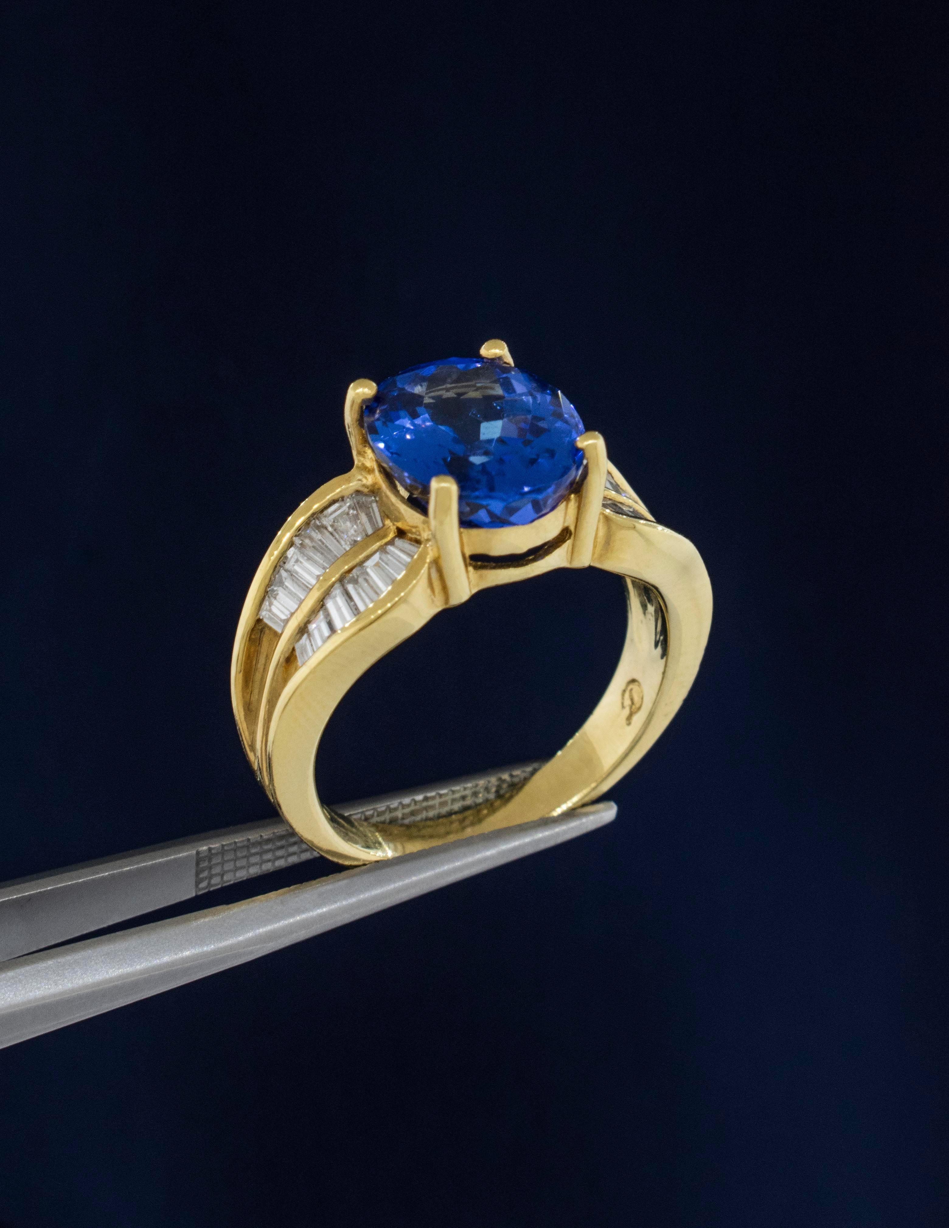 An 18K Yellow Gold Tanzanite Ring with Baguette Diamonds.
The Oval Tanzanite is an Ideal Color for  the mineral Zoisite. It is a deep lavender blue weighing approximately 3.22 carats, grading VS Clarity for a Type I stone. 
The Diamonds are Baguette