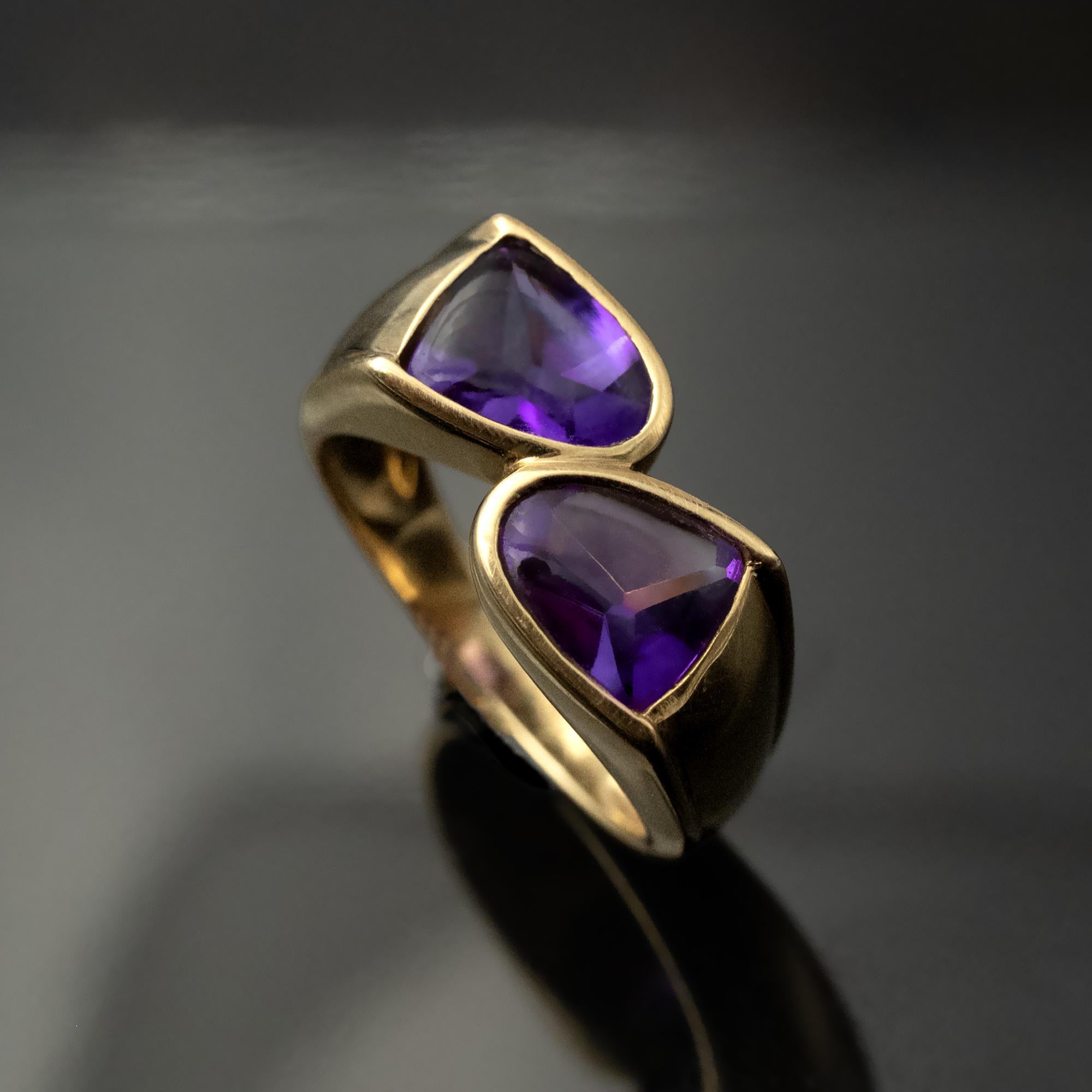 Cette bague toi et moi en or jaune 18 carats, proposée par Claris.A, présente deux améthystes positionnées sur un anneau ouvert, se faisant face dans une composition nette et équilibrée. Les deux pierres sont taillées dans un style buff-top lisse,