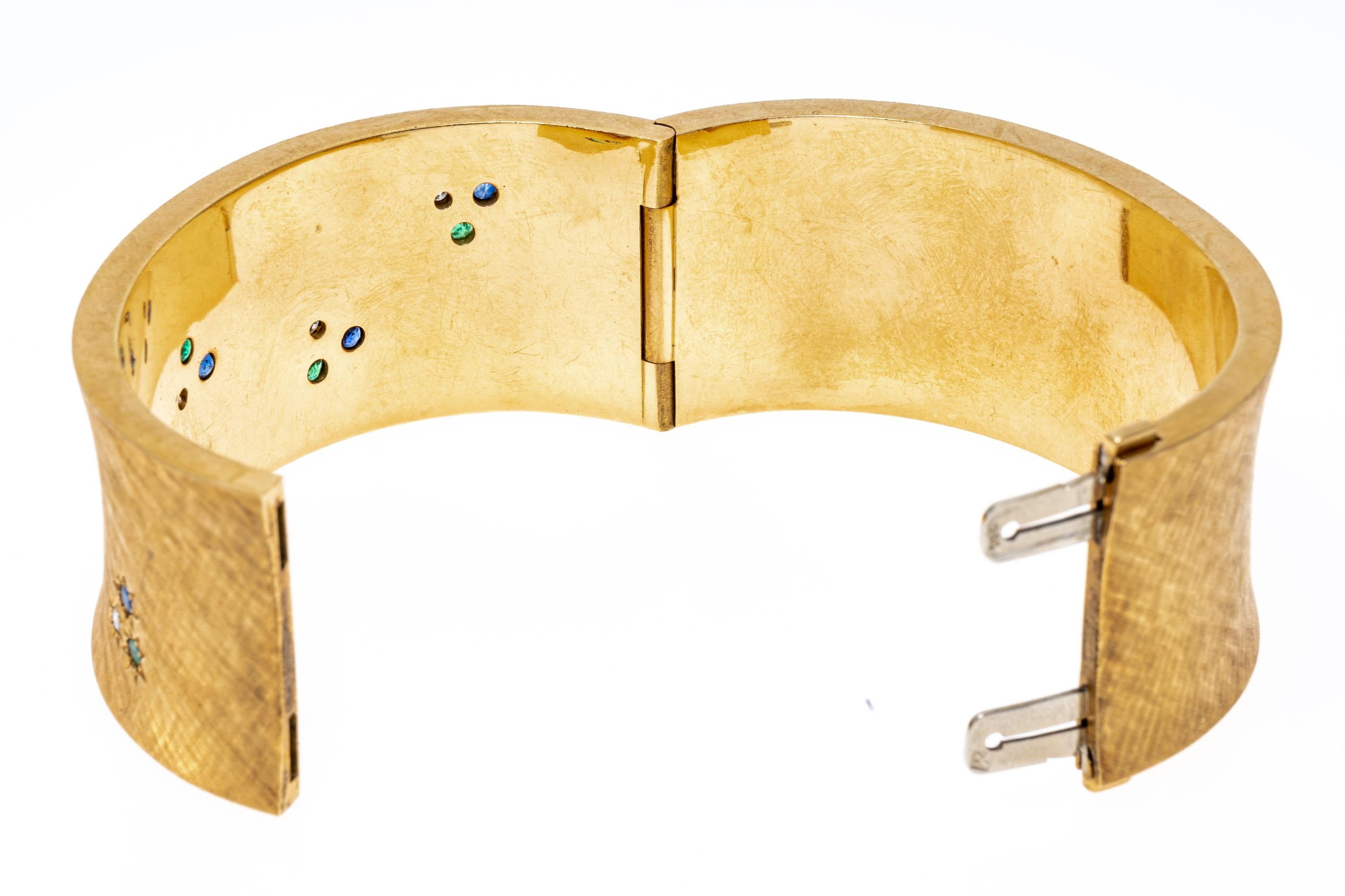 Fabriqué en or jaune 18 carats, ce large bracelet concave présente une douce finition brossée. Des émeraudes, des saphirs et des diamants de taille ronde sont sertis à fleur de la face du bracelet dans un cadre en forme de soleil. Les diamants ont