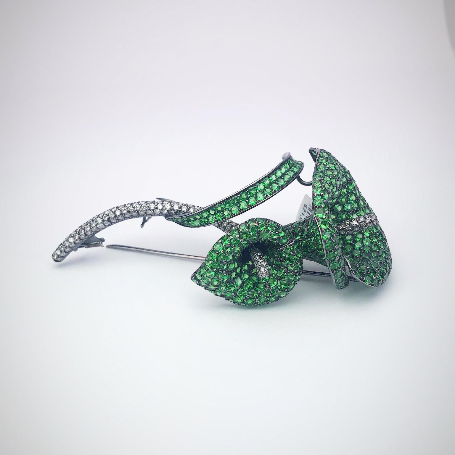 A three dimensional calla lily brooch. The two flowers are set entirely with green garnets and round brilliant diamonds. The stones are set in blackened gold for a dramatic effect. The flowers measure 3.75