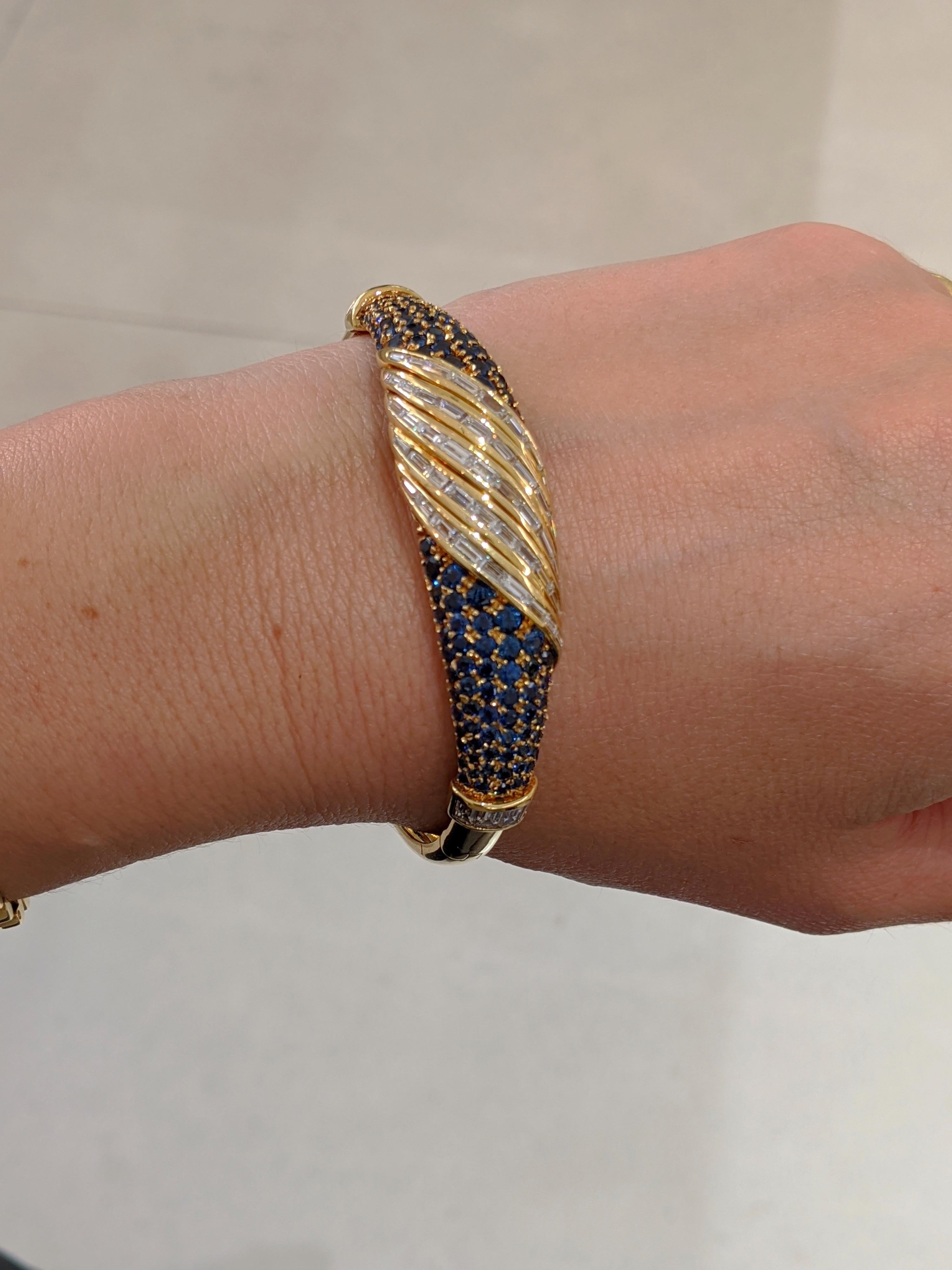 An amazing retro bangle bracelet.  The center swirl section of this 18 karat yellow gold bangle has been invisibly set with Baguette Cut Diamonds. The two side sections are set with round  Brilliant Cut Blue sapphires. The inside measurements of the