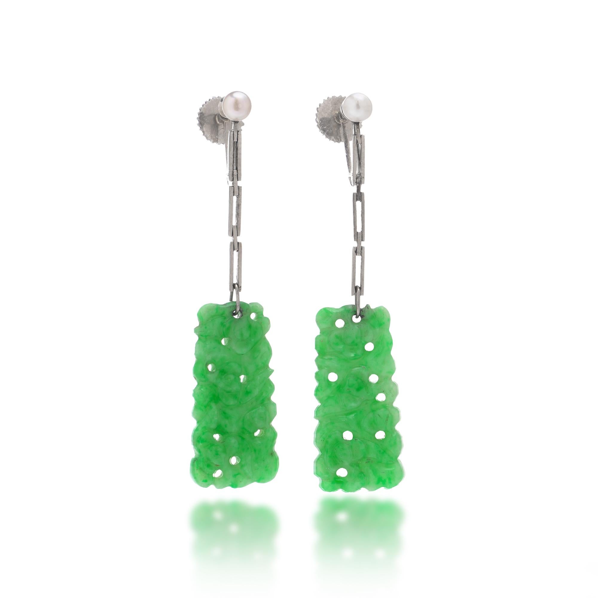 Une délicate paire de boucles d'oreilles en or 18kt avec des pendentifs en jade finement sculptés, chacun représentant des motifs floraux stylisés dans de riches tons verts. Les gouttes de jade sont suspendues à des fermoirs à vis sertis de petites